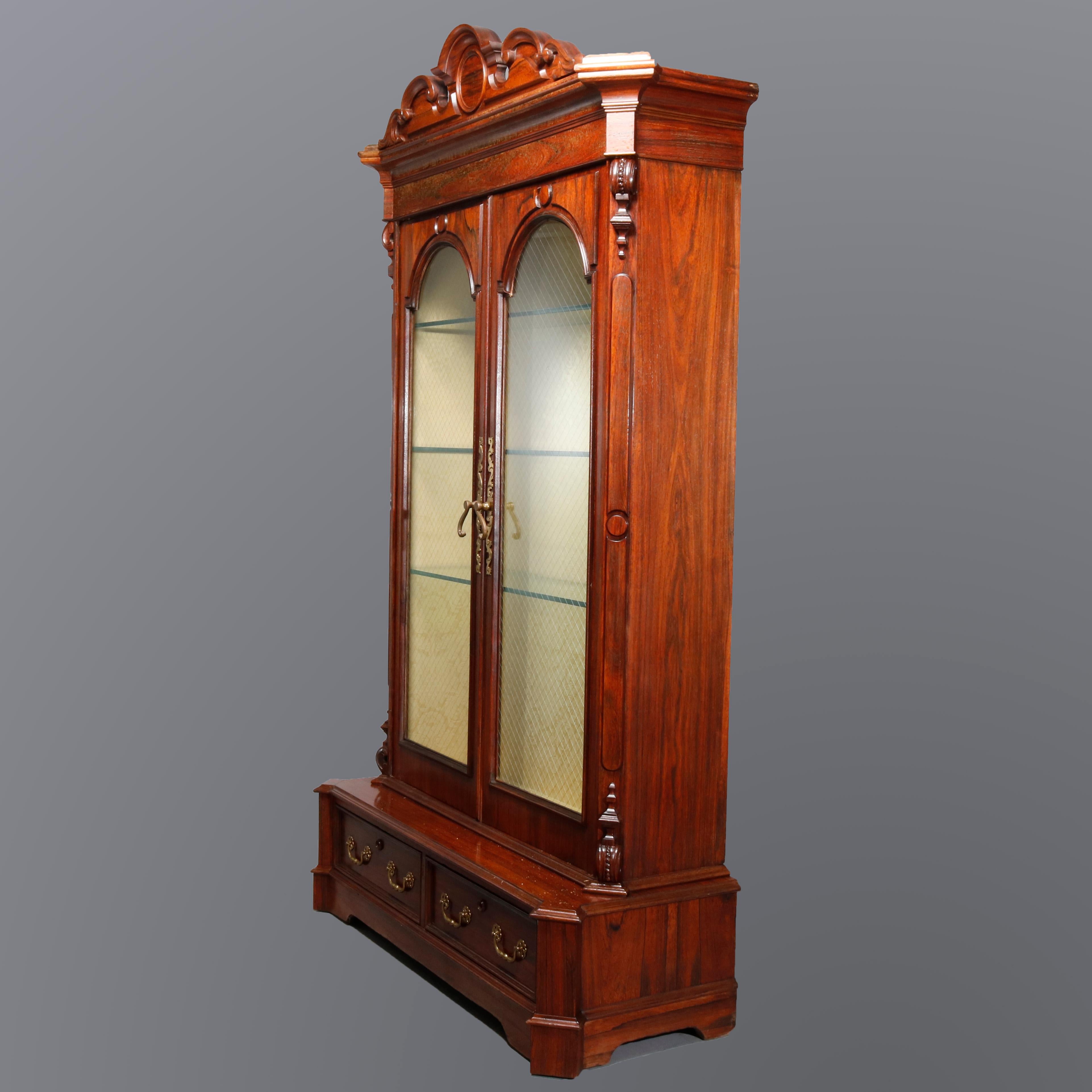 An antique Victorian china display cabinet in the manner of Joseph Meeks offers rosewood construction with scroll form crest surmounting double doors with arched glass opening to lighted case having glass shelving, raised on base with two drawers,