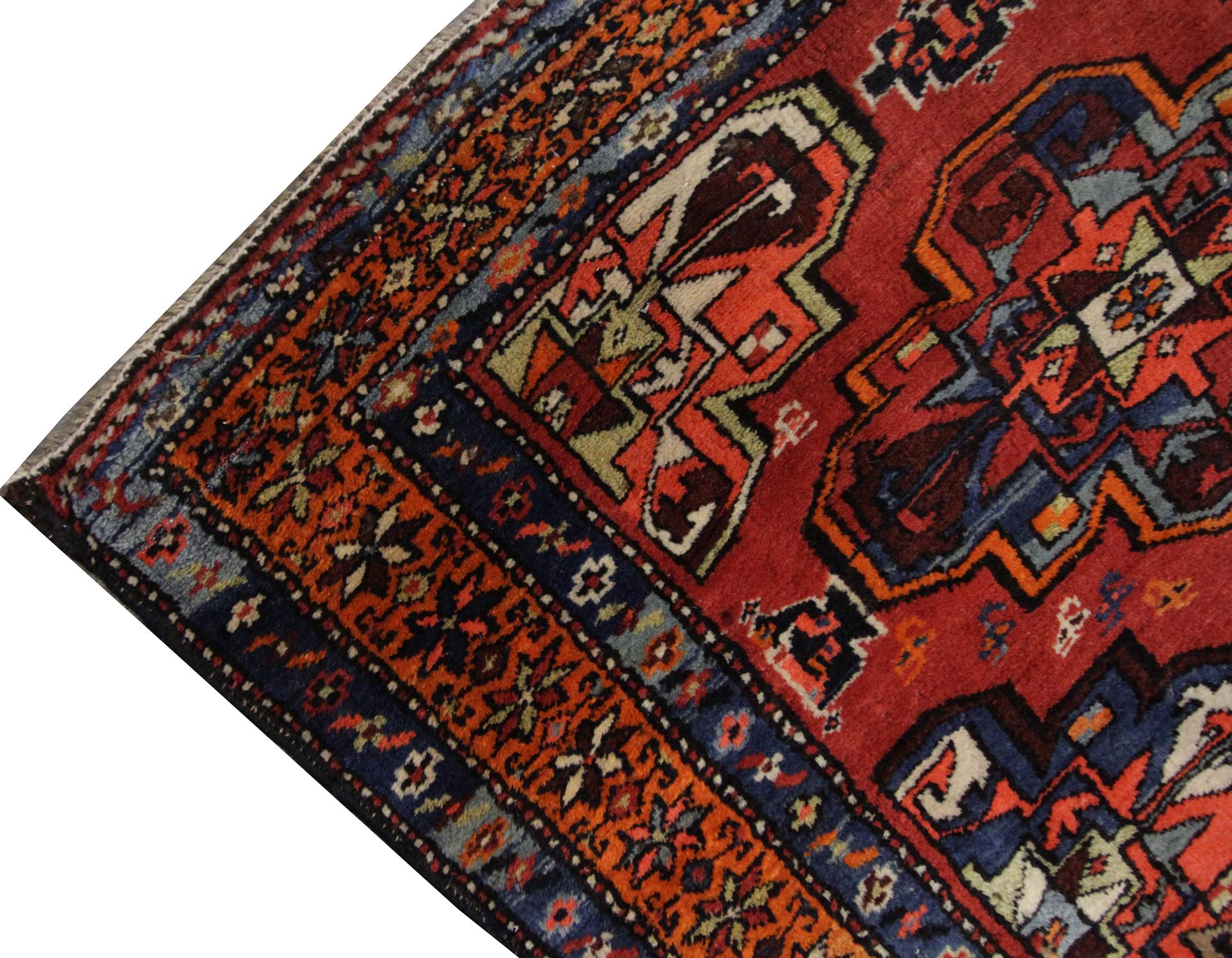 Un tapis fait à la main avec un motif répétitif de dessins géométriques coule au centre de ce tapis oriental, avec des couleurs vertes, bleues et brunes contrastant doucement avec le fond orange foncé, entouré d'une bordure à motif répétitif. Riche