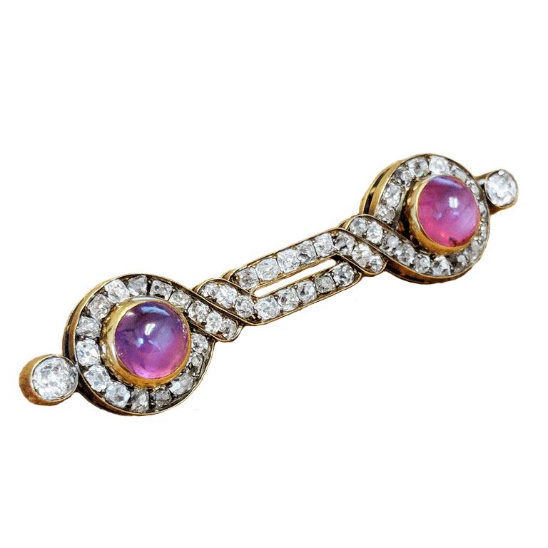 Designed as a stylised infinity symbol, this antique brooch is set with old mine cut diamonds for a total weight of approximately 3 carats. At each end is a purplish-pink star sapphire. The brooch is mounted in silver and 18 karat yellow gold. It
