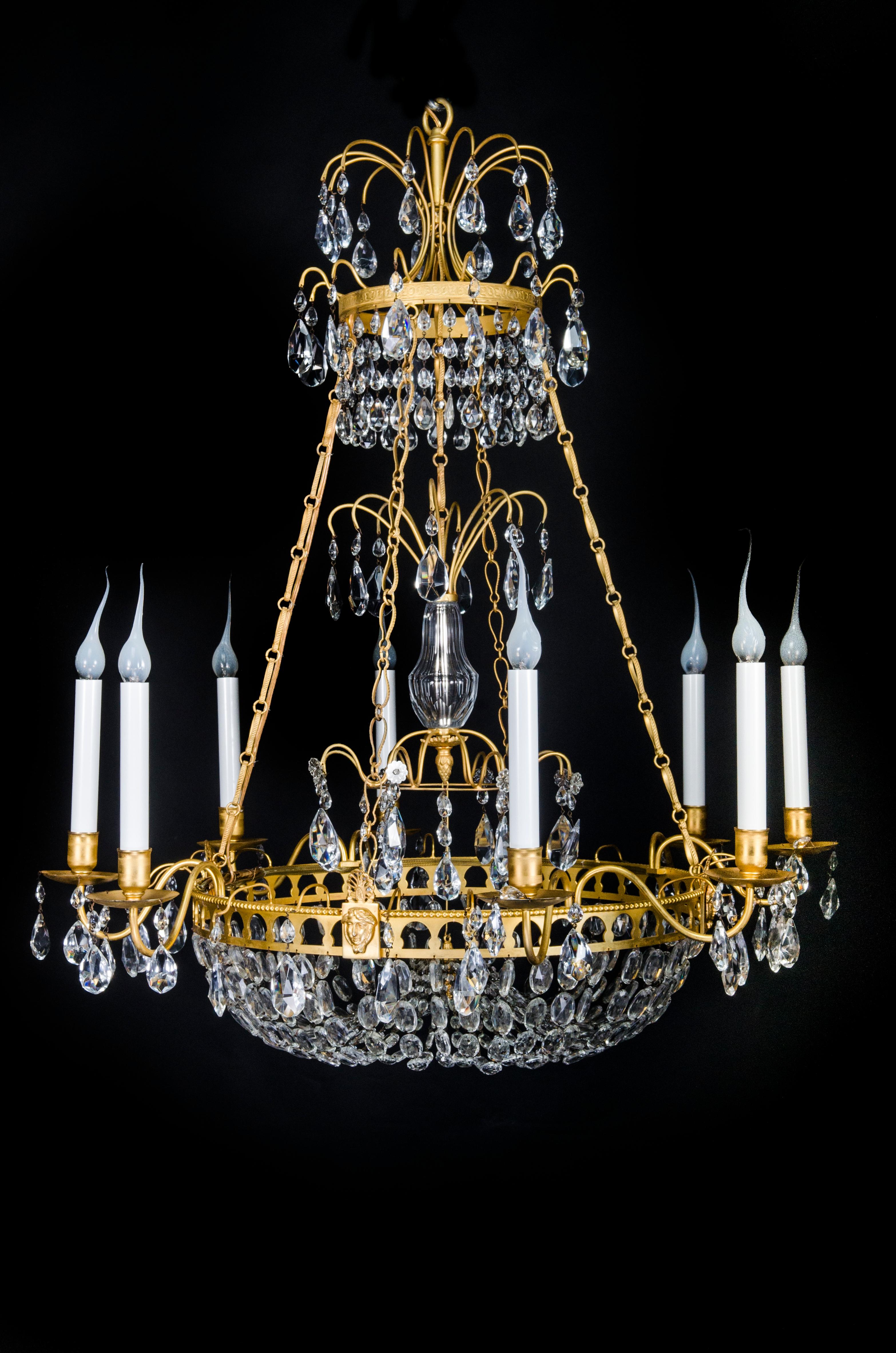 This antique Russian Neoclassical chandelier features a triple-tiered gilt bronze frame adorned with cut crystal prisms. The structure is composed of a circular lower tier with eight arms, each fitted with candle-shaped holders. The arms extend from