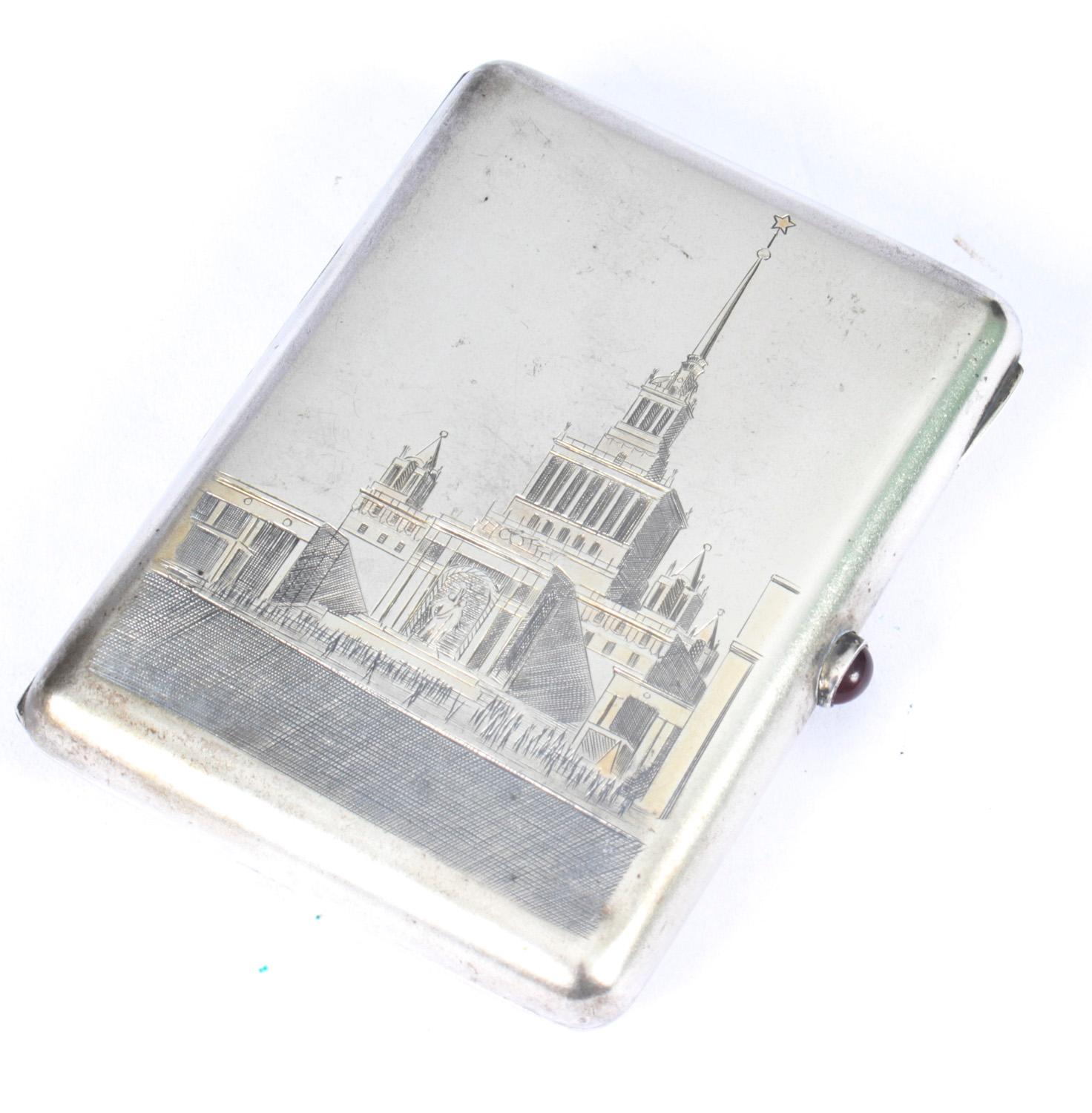 This is an exquisite mid 20th century Russian silver and niello rounded rectangular cigarette case, the cover decorated with Moscow State University, the Communist skyscraper surmounted by a Soviet star, with a ruby cabochon push-button mechanism,