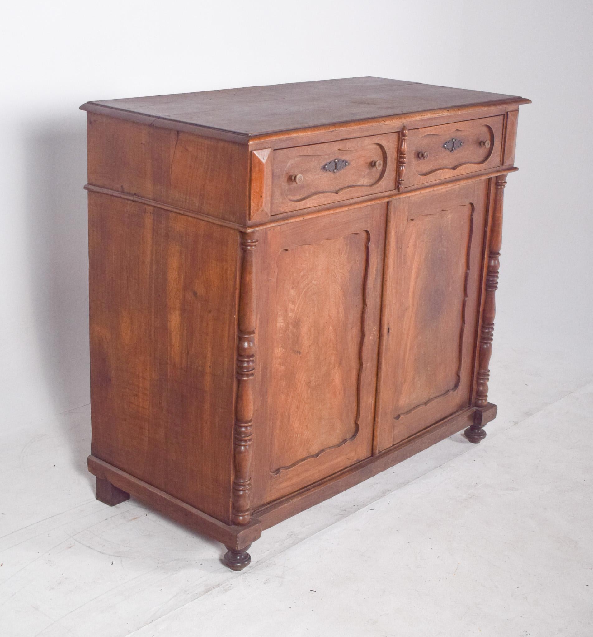 Rustic wardrobe with two doors and two drawers. With colonial and classic style features, the wood has a natural finish, with a warm colour and visible veins. The drawers have rounded handles and carved details, as well as decorative metal plates