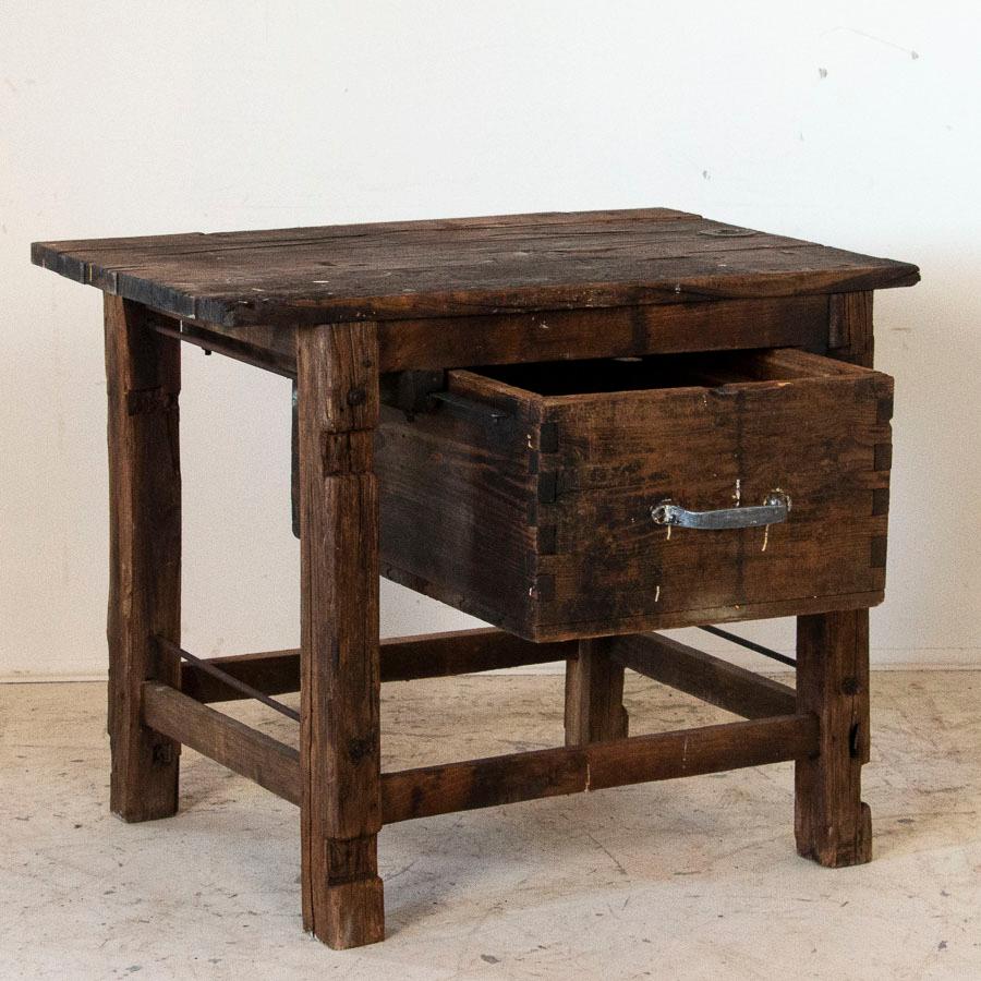This primitive work table reveals its age in every element, from the 6 planks of wood forming the top to the heavy metal bolts and rods and thick dove-tail joints. It is in rustic, well used worn condition with paint and oil stains reminding one