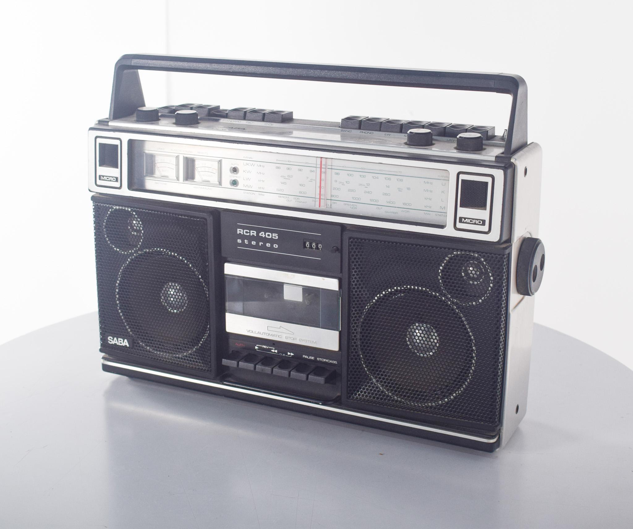 Radio-cassette portable SABA RCR 405. L'appareil présente un design robuste et fonctionnel, avec une structure rectangulaire dans les tons noir et argent, accompagnée d'une poignée supérieure pour faciliter le transport.
La face avant comporte un