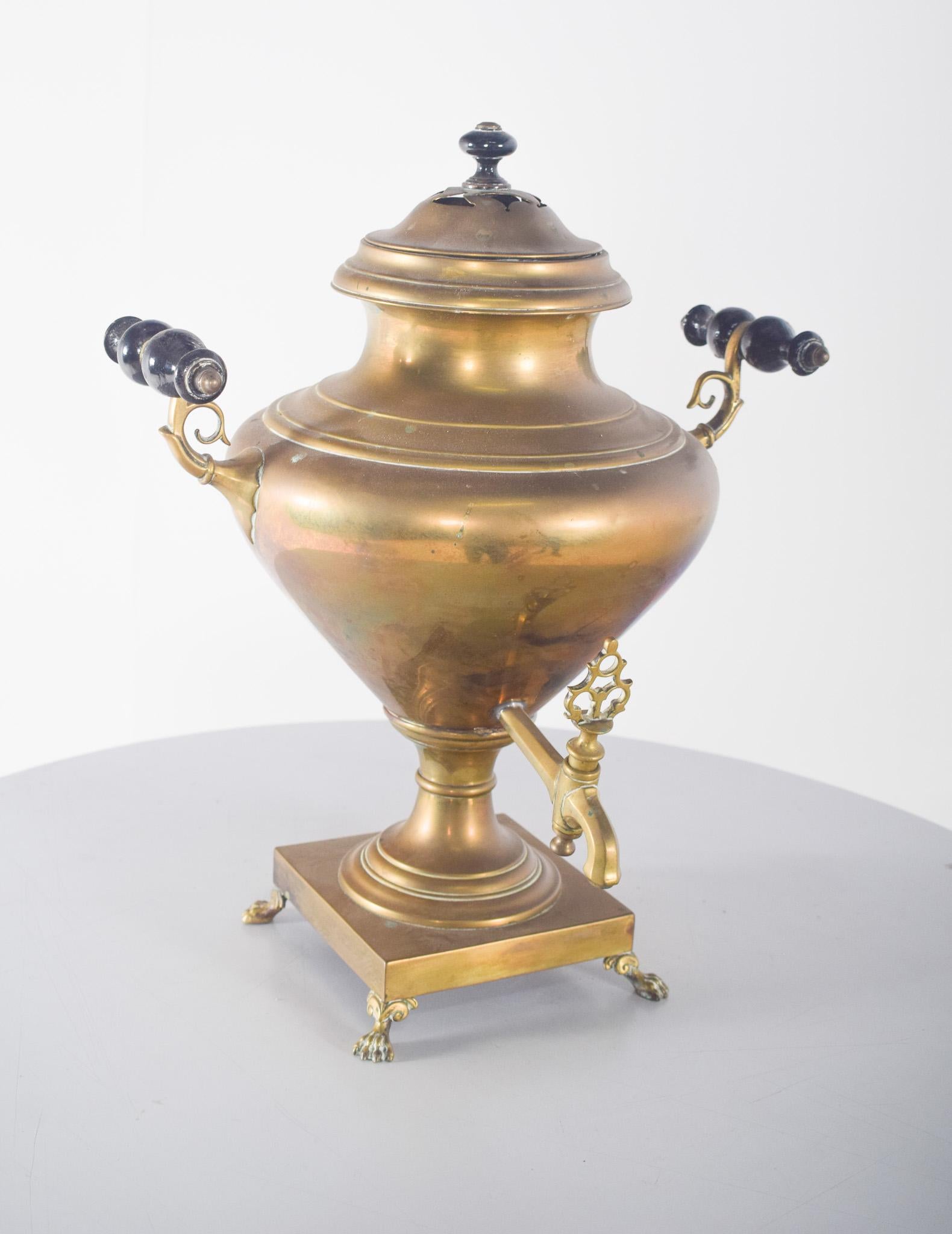 Samovar en laiton jaune. Avec une structure robuste, un corps bulbeux et une finition vieillie qui révèle une patine caractéristique du temps. Il possède une base carrée avec des pieds en forme de griffe qui offrent stabilité et élégance.
Les