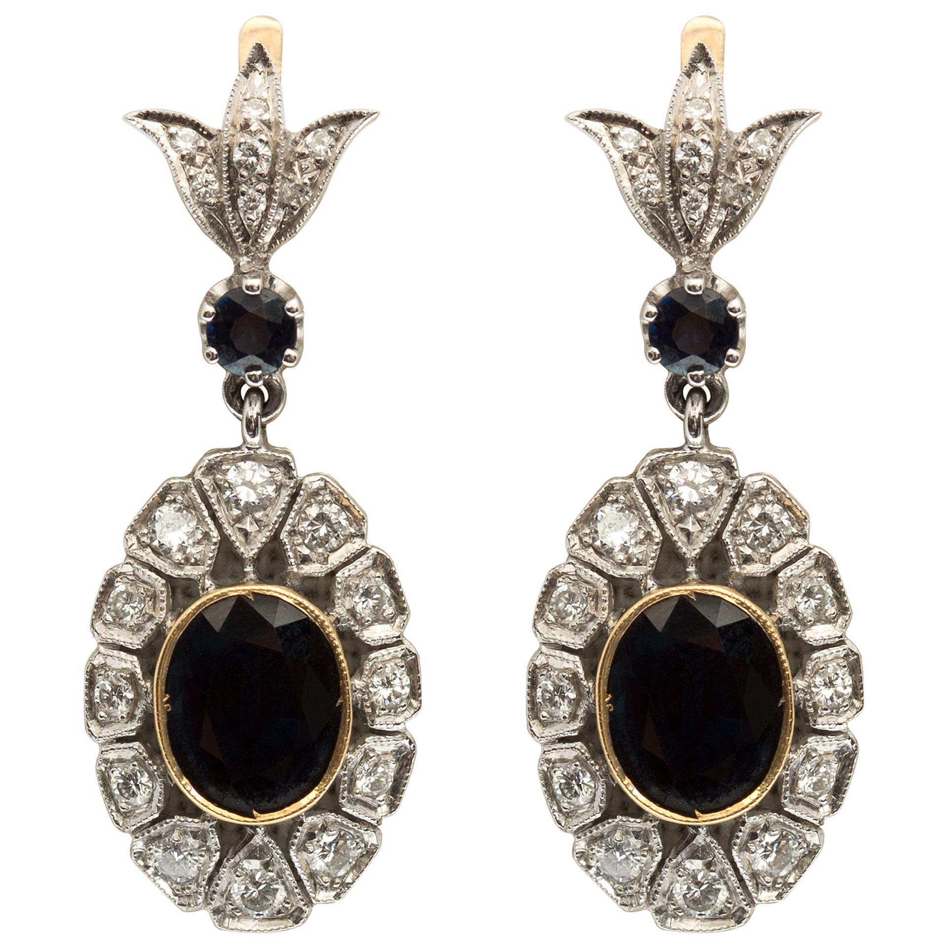 These elegant earrings feature two oval bezel set sapphire drops framed by stylized petals of bead set round diamonds each surmounted by a single round sapphire and a delicate foliate design diamond top. Measuring 1 ¾” and completed by hinged back