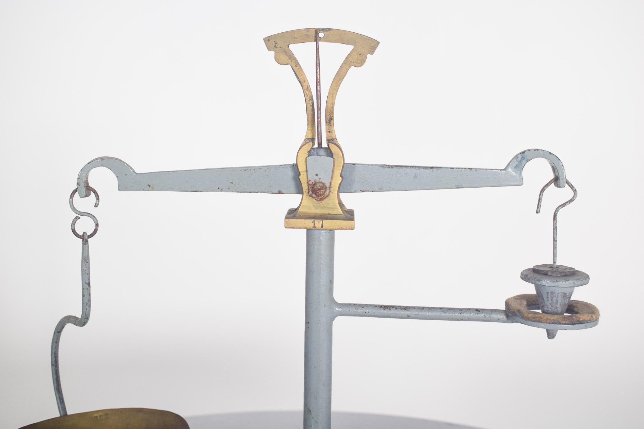 Balance ancienne en forme de croix avec un plateau et un poids. Structure principale peinte en gris, avec des signes d'usure qui soulignent son caractère historique. Le mécanisme du balancier est constitué d'une tige horizontale surmontée d'un