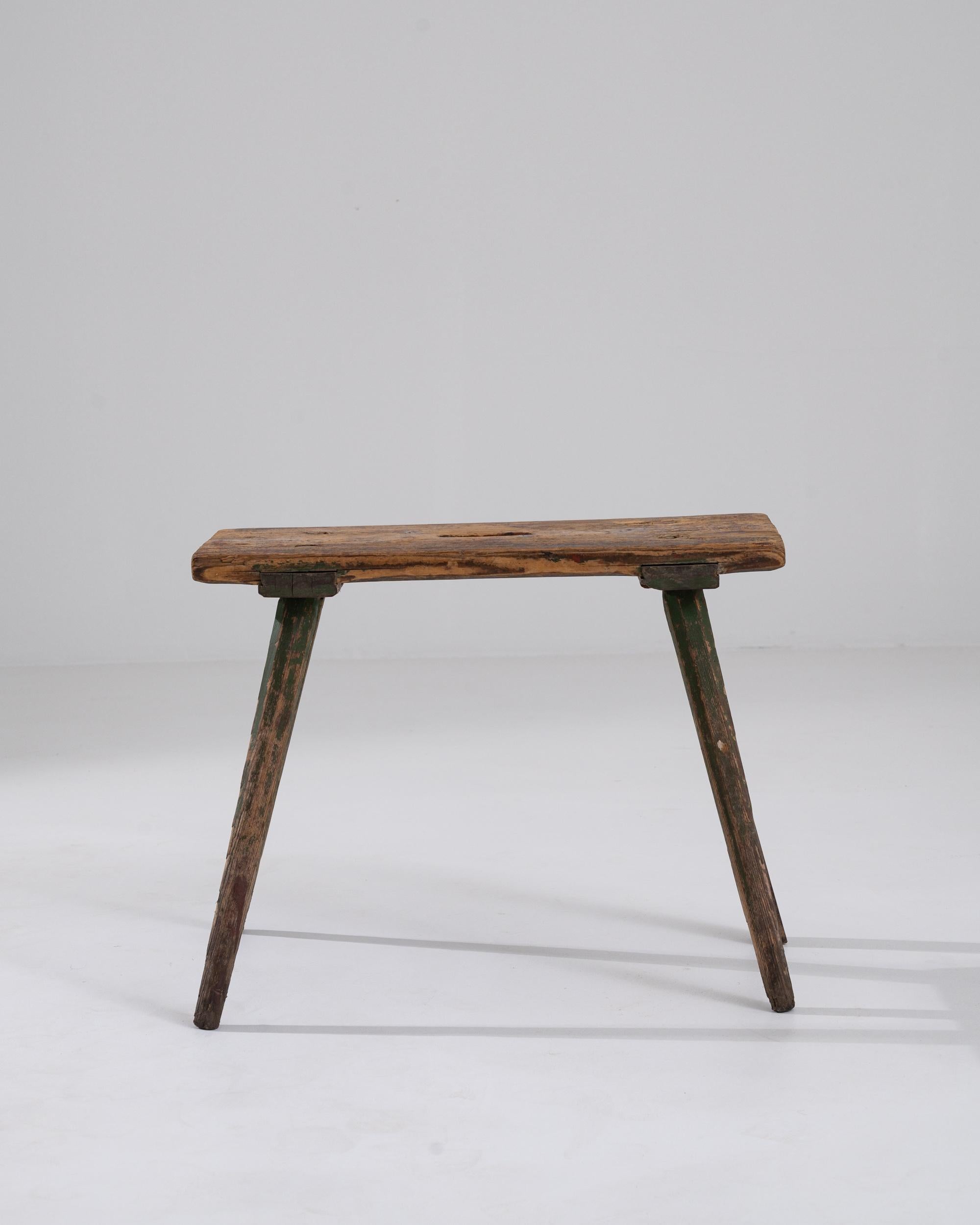 A wooden stool made in 19th century Scandinavia. A simple wooden stool composed of one seat plank and four diagonal legs, all of which have been slowly covered in an earthy patina. The minimalist image of the stool’s form conjures a picture of a