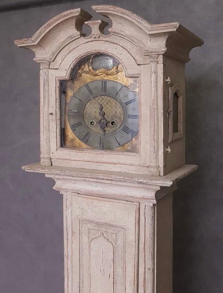 This beautiful beige colored Danish clock with rich decorations was produced circa 1800 and comes from Bornholm. The upper part has a larger rich carved finial and features glass-side windows on the sides. The clock features subtle carvings on the