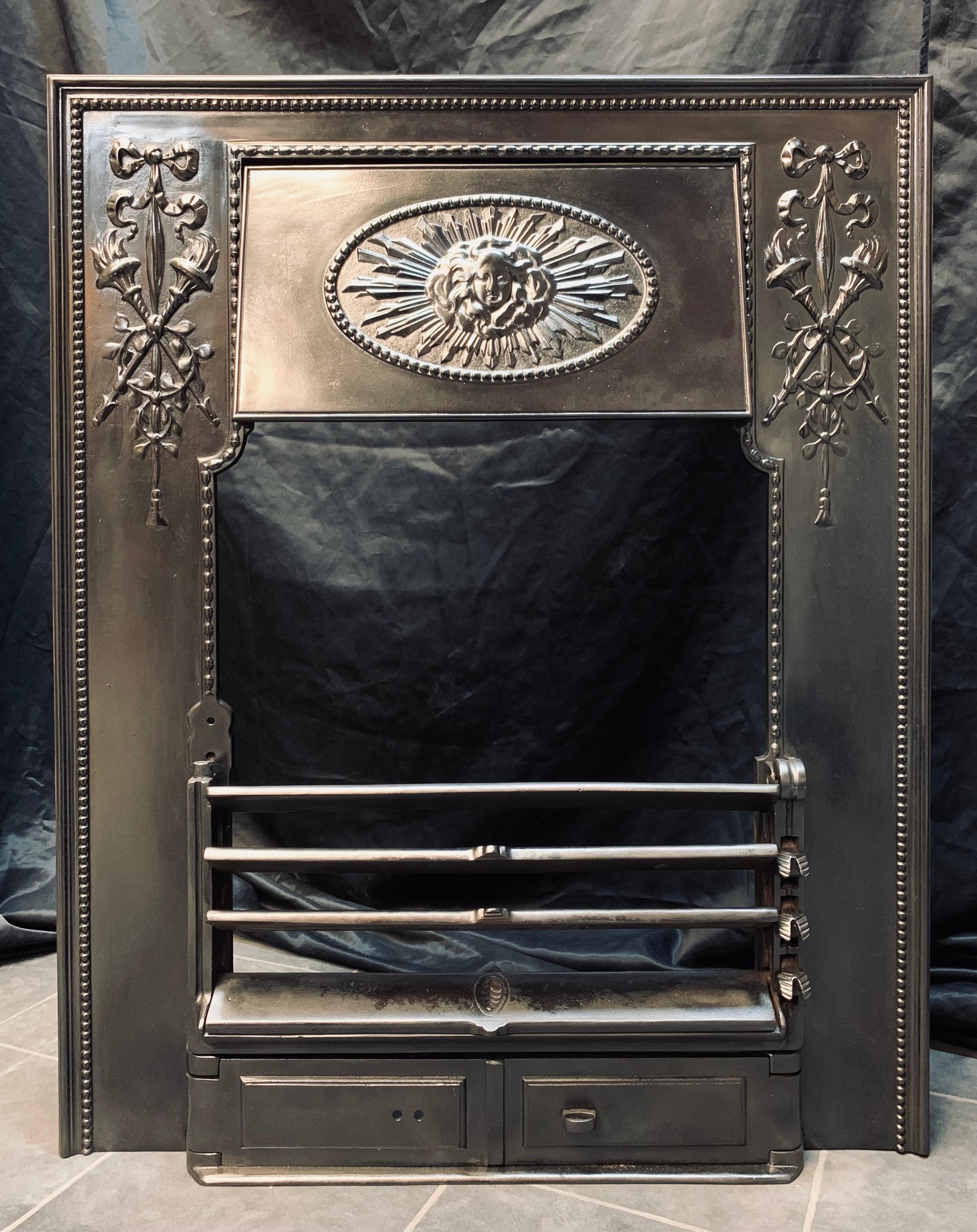 A well cast antique Scottish cast iron fireplace insert probably by Carron of Falkirk. A framed and beaded border with panels of tied ribbon and tassel with crossed flaming torches, a central movable hood set in an oval beaded border with a mask
