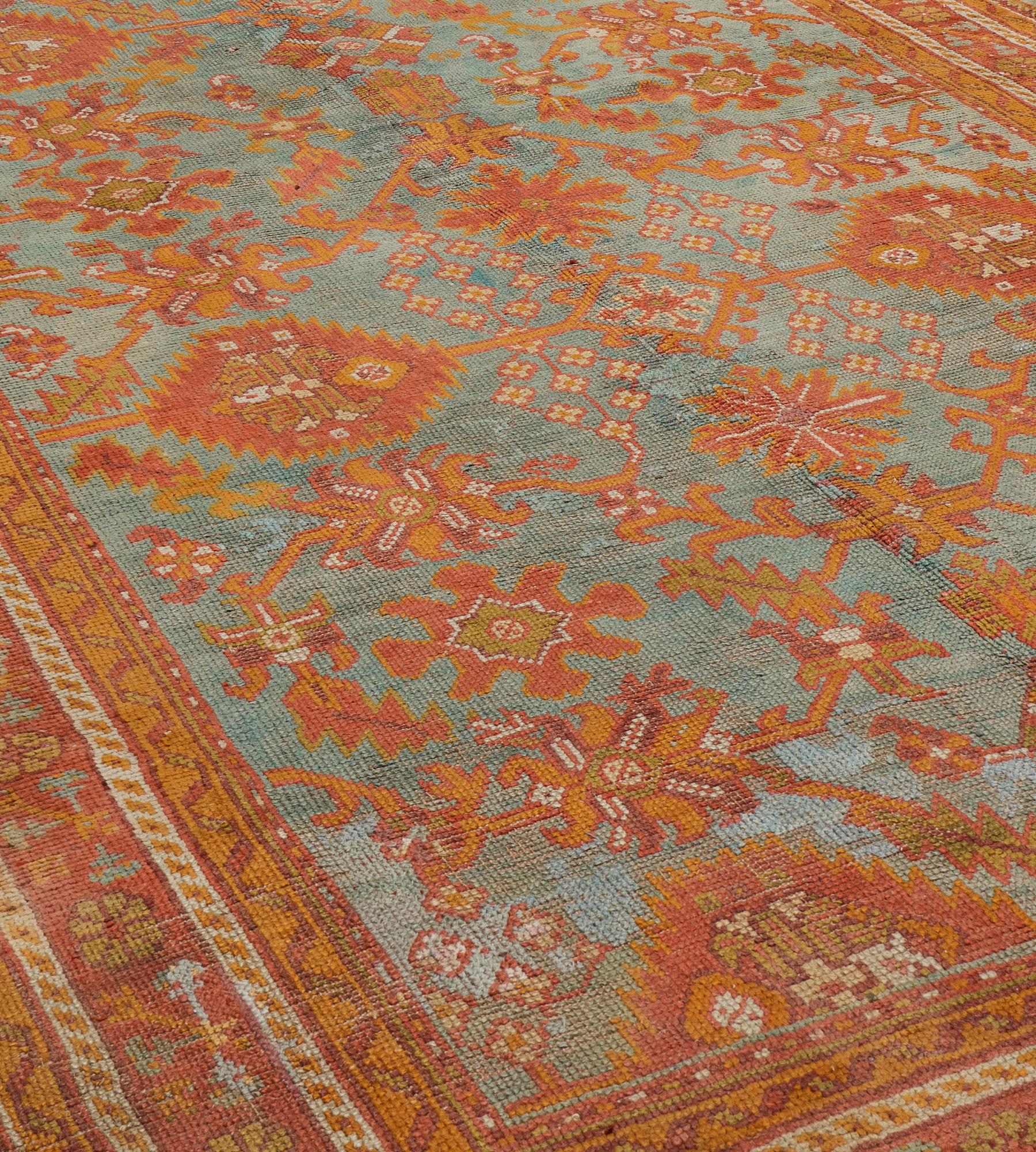 This antique Oushak rug has a sea-blue field with an overall design of tomato-red, burnt-orange bold angular palmette and floral leafy vine, in a tomato-red border of camel-brown rosette and floral vine between ivory panel and burnt-orange ribbon