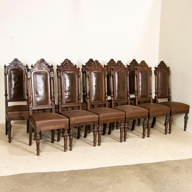 This striking set of 12 tall dining chairs are made dramatic due to the carving and finials that embellish the upper top, turned column sides along with attractive turned legs. The seats and backs are brown leather with nailhead trim. Note the