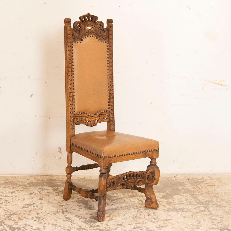 This striking set of 12 tall dining chairs are made even more dramatic due to the carved King's crowns that embellish the upper top and lower stretcher, along with carved feet as well. The seats and backs are tan leather and appear to be relatively