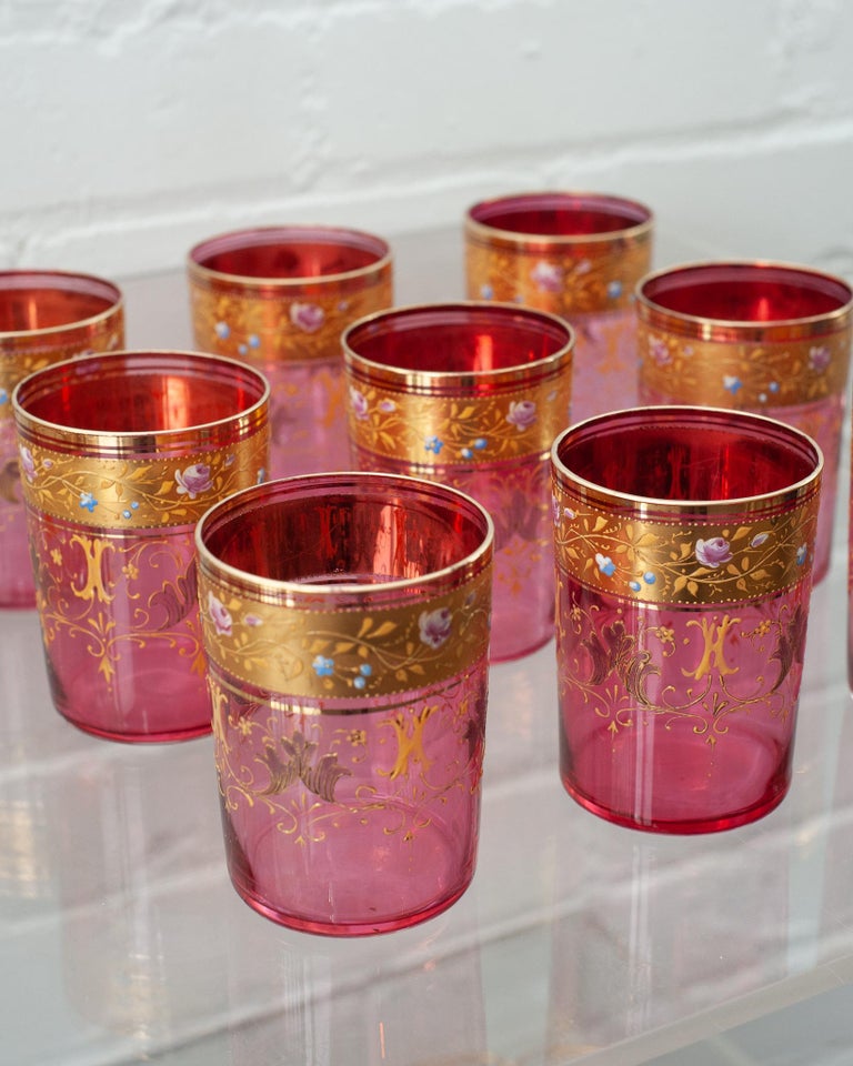An usual and delicate set of 9 Antique Moser cranberry & gold gilt tumbler glasses. This thin cranberry green glass is gilded ornately in gold scroll motifs.
 