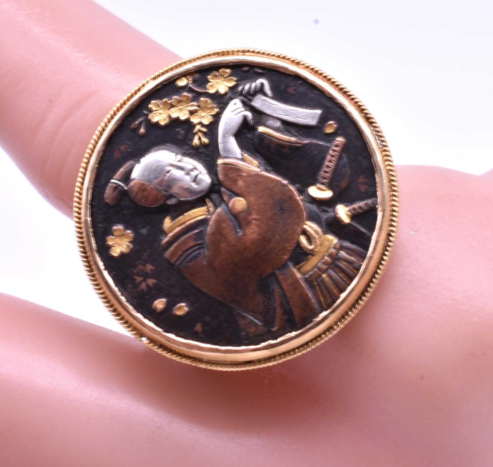Wir bewundern diesen Shakudo-Ring aus dem Jahr 1880, der einen japanischen Samurai-Krieger mit zwei Schwertern in traditioneller Kleidung zeigt, der seine Wünsche auf Papierstreifen schreibt, die er an Bambuszweige hängt. Nach japanischer Tradition