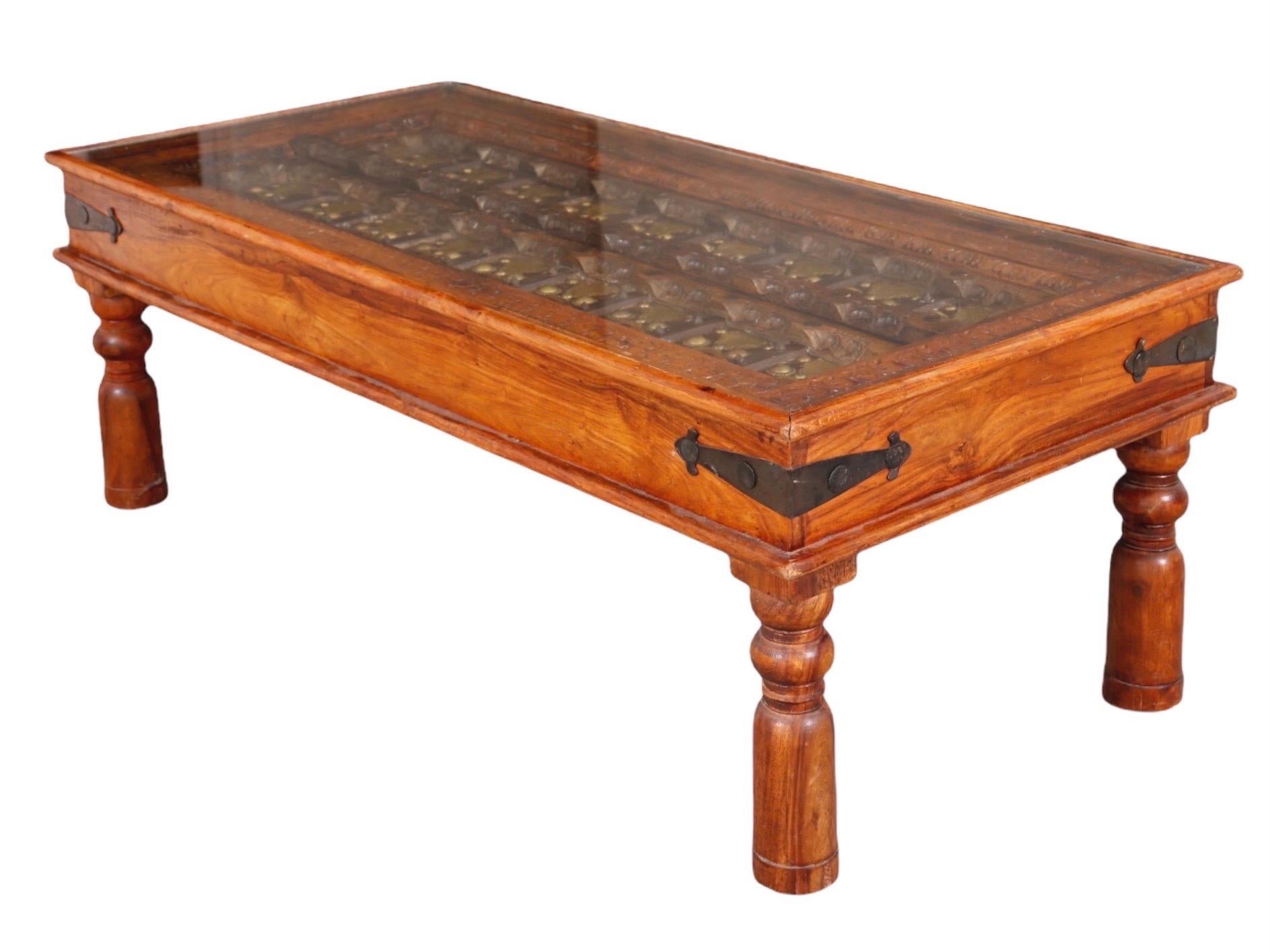 A glass top coffee table made with an antique Indian embellished sheesham wooden door. Dating from the late 19th century, the door panel consists of elaborately carved sheesham wood decorated with brass and iron studded bands. Framed in knotty pine,