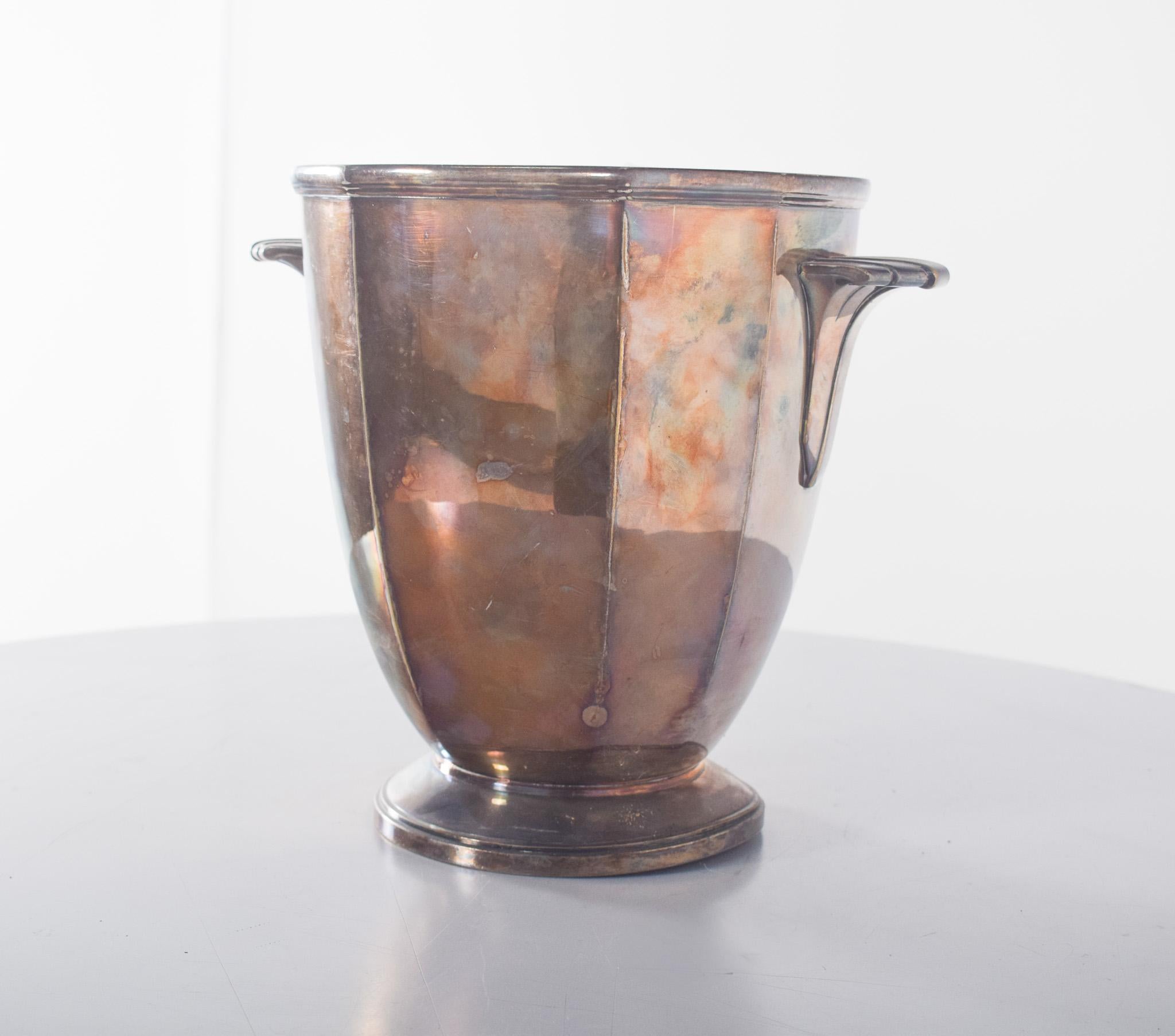 Shell ice bucket. With a slightly conical body, widening at the top and resting on a rounded, firm base.
It has two discreetly integrated side handles, which facilitate handling. The outer surface shows oxidisation and patina marks, the result of