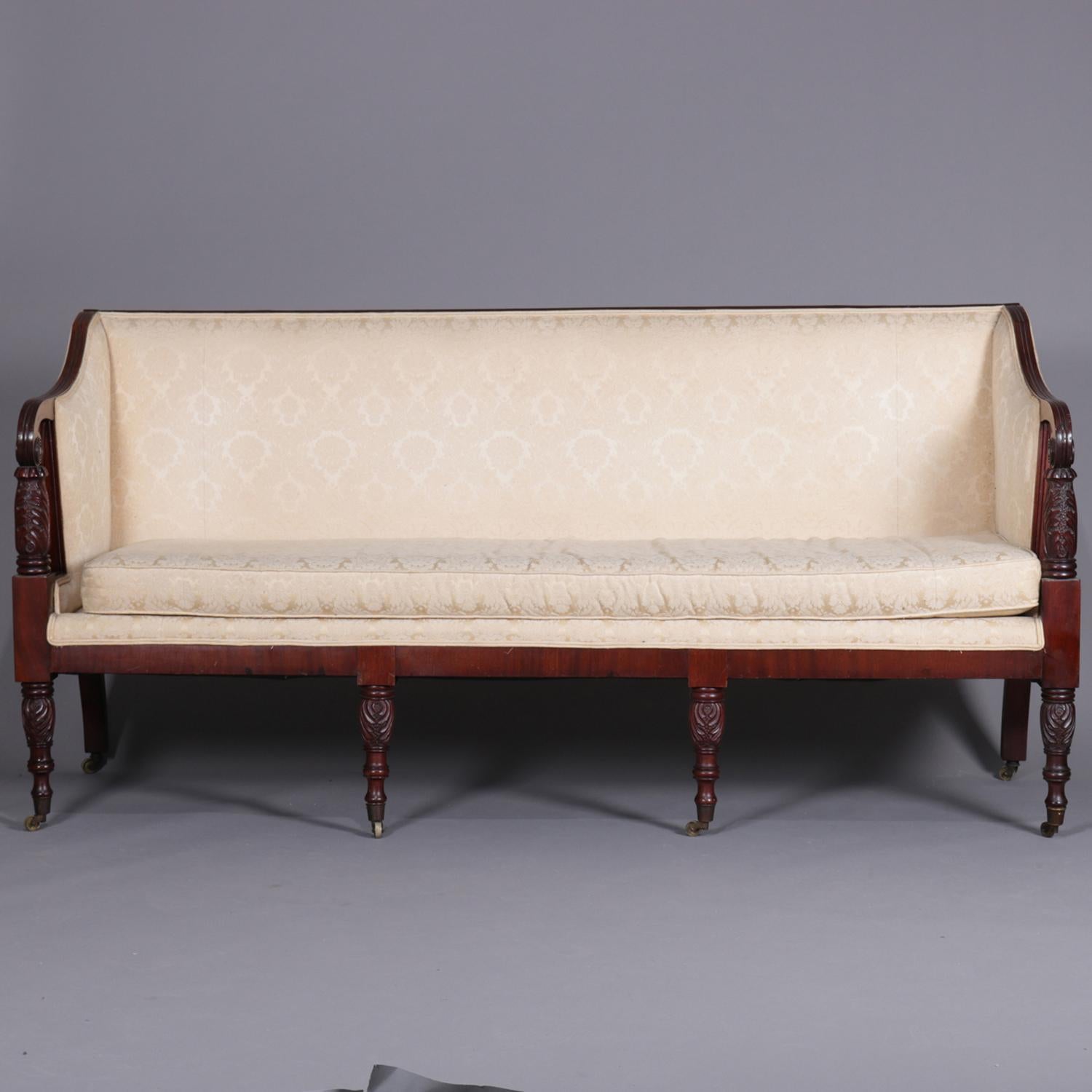 An antique period Sheraton classical Samuel McIntire School sofa features mahogany frame with scroll form open arms having central rosettes over foliate carved columns, raised on turned and foliate carved legs terminating in casters, newer