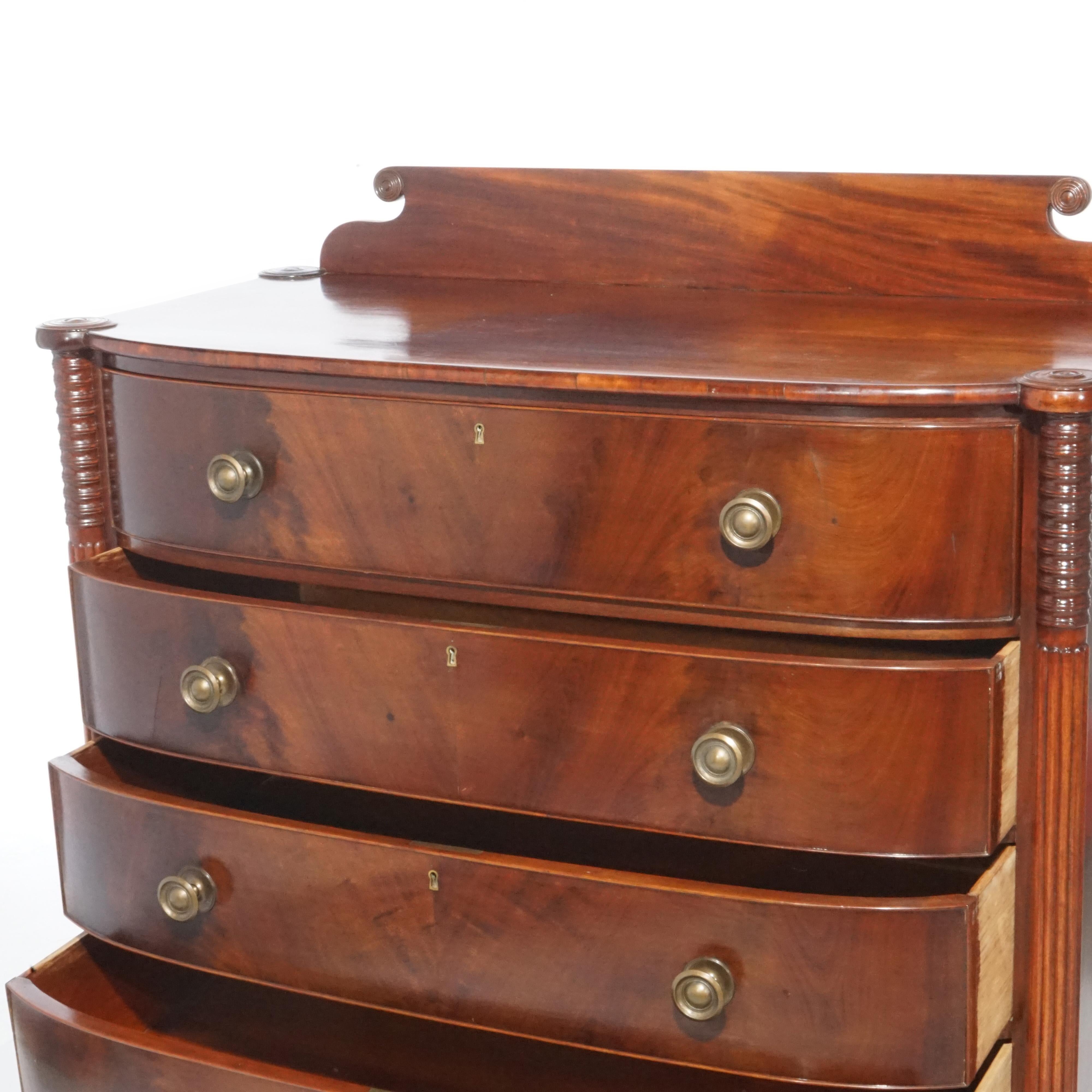 An antique Sheraton chest of drawers offers flame mahogany construction with scrolled backsplash over swell front case having four bookmatched long drawers flanked by reeded columns and raised on turned legs, c1820

Measures- 47'' H x 46.25'' W x
