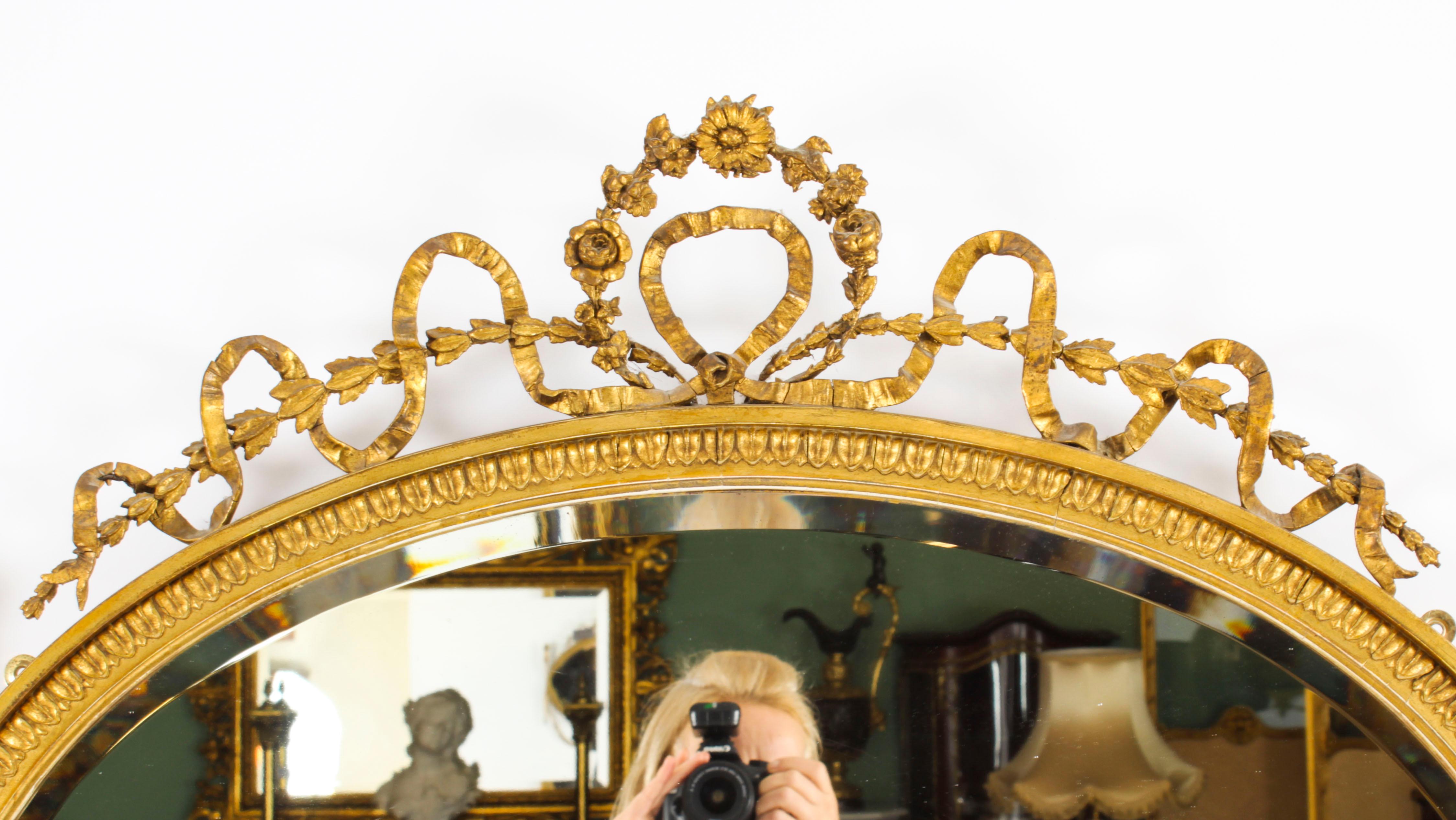 This is an exquisite antique Sheraton Revival oval hand carved giltwood mirror, circa 1860 in date.
 
The mirror features an oval bevelled mirror plate in a gadrooned frame decorated with trailing ribbons, blue bell flowers and flaming torches to