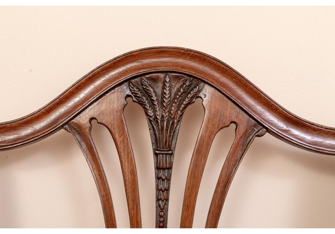 Finely carved three-seat shield backs with a darker carved wheat sheaf motif at top of the curving openwork splats. The arms with carved bell flowers. Raised on front cabriole legs with acanthus leaf carved and scrolled knees and ball- and- claw