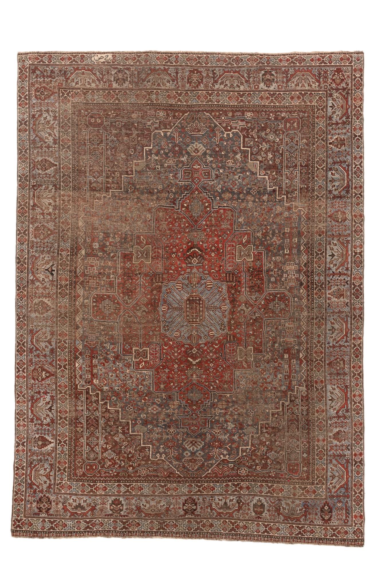 Un sorprendente tappeto antico Shiraz, proveniente dalla Persia sud-occidentale, che mostra un audace medaglione centrale in un profondo rosso mattone circondato da forme geometriche a gradini in blu e taupe. Il campo è riccamente ornato da rosette