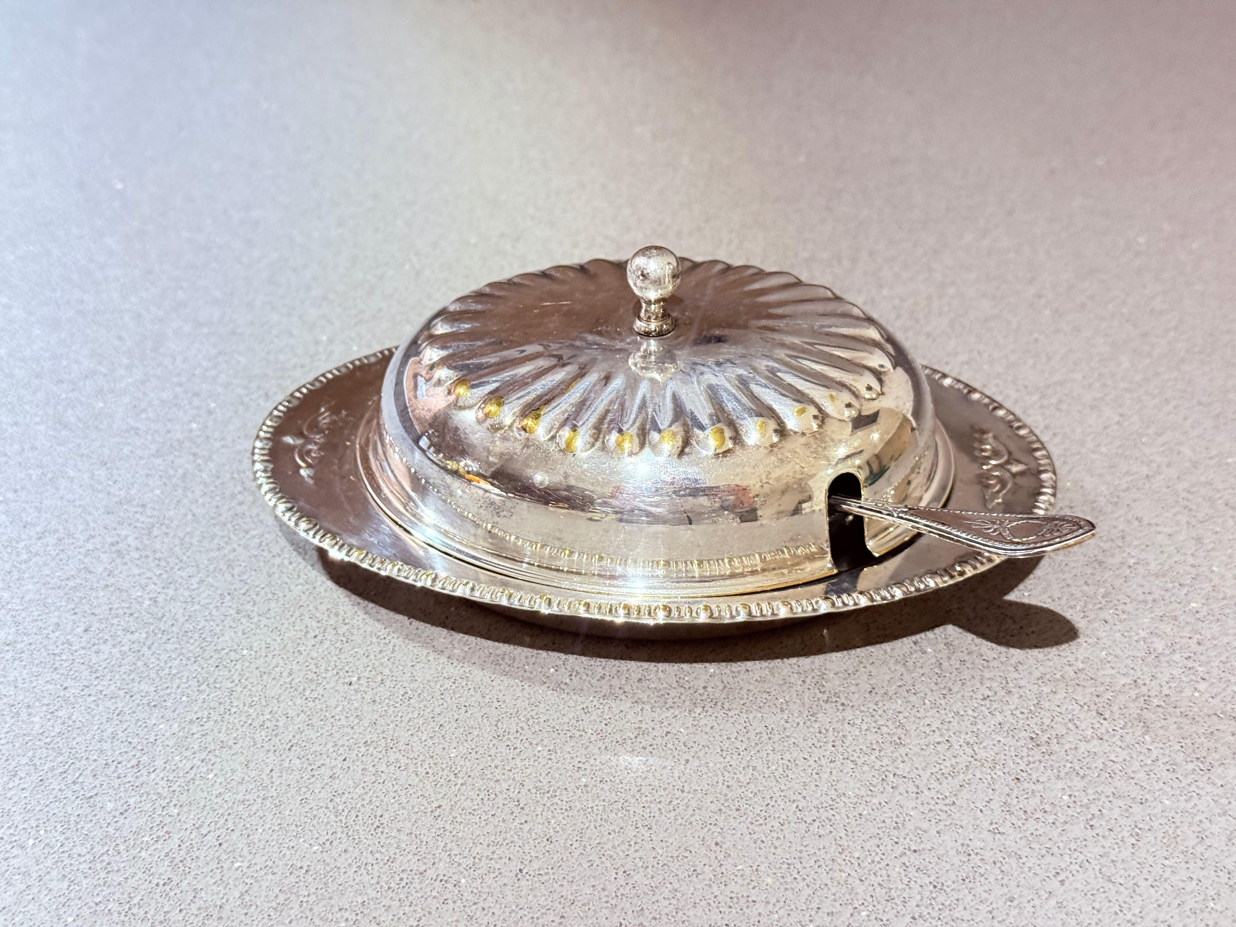 Beurrier ancien en argent avec couvercle en cristal et cuillère, vaisselle vintage élégante
Ce beurrier en argent ancien est un exemple frappant d'élégance vintage, avec un revêtement en cristal présentant un motif classique de rayons de soleil et