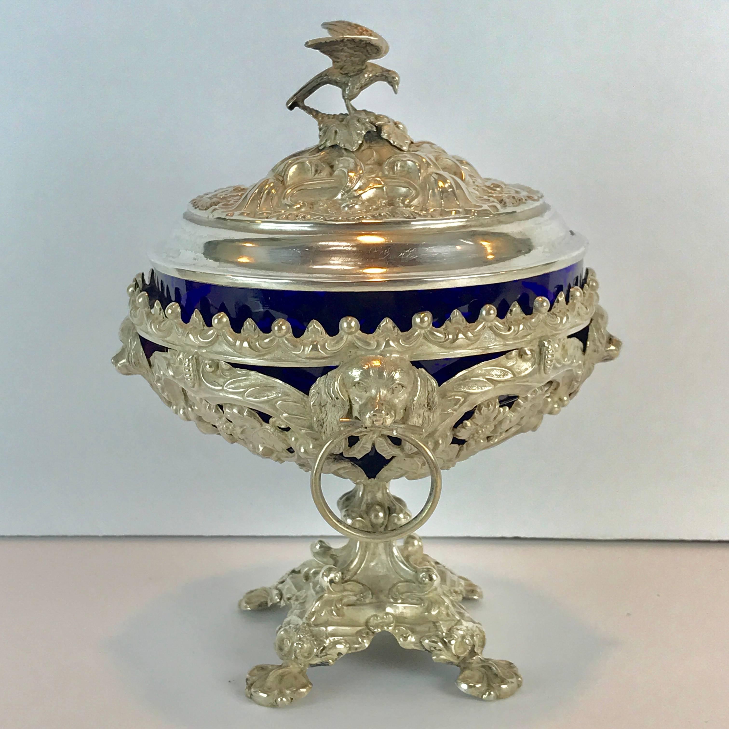 Antique silver dog motif covered cobalt urn, the reposed lid with eagle finial, the body with cobalt blue glass liner in a pierced body with four dog motif medallions, raised on a paw footed pedestal base. Both the lid and base are marked with