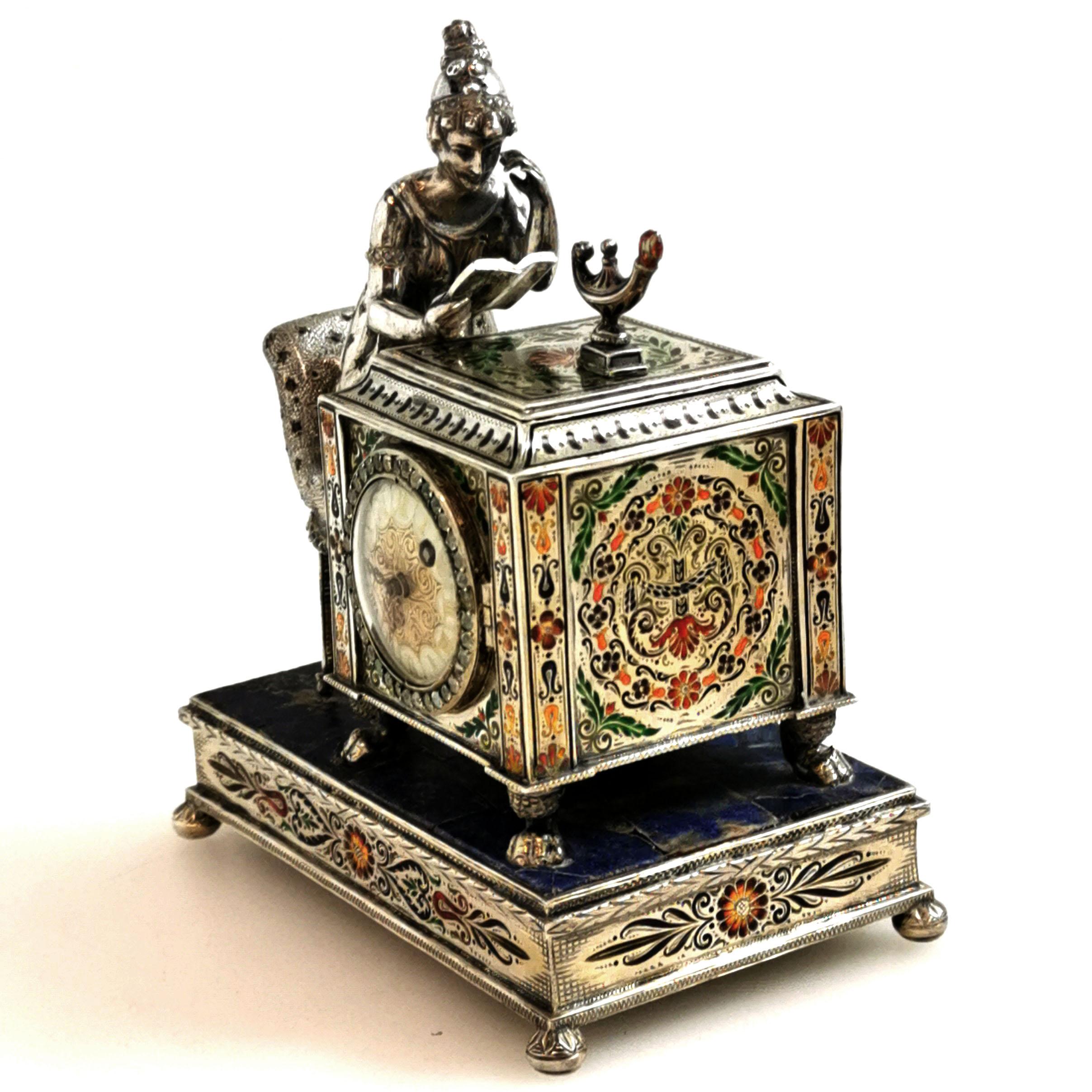 A beautiful Antique Silver and Enamel table reading Clock inset with rubies, sapphires and other gems. The Clock is in the form of a lady in period dress sitting at a desk reading by the light of an oil lamp. The Clock has a silver mounted Lapis
