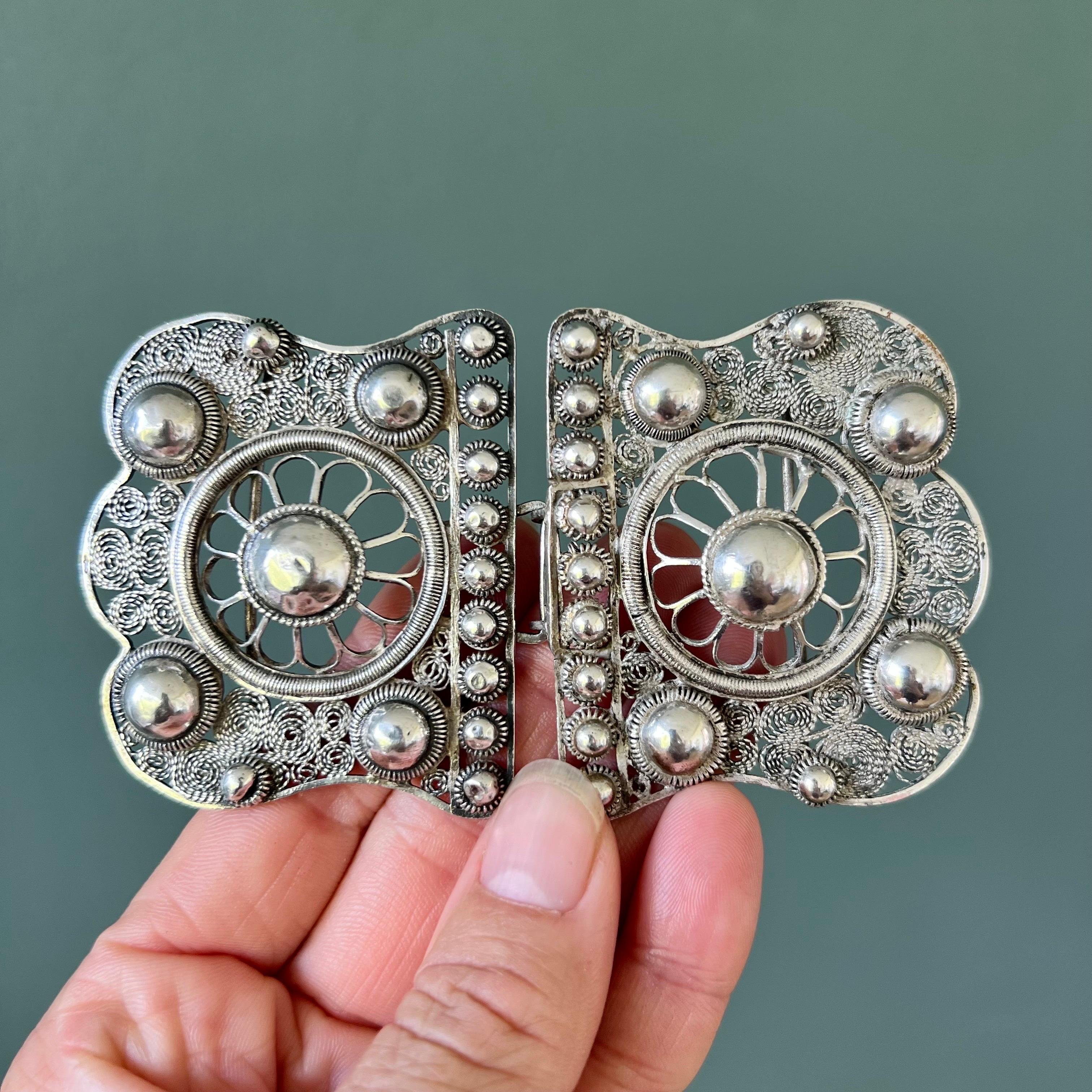 This antique silver ornate floral filigree belt buckle is a beautiful example of intricate craftsmanship. The buckle features a striking design centered around two prominent silver spheres, one set on each side. These spheres are encircled by a
