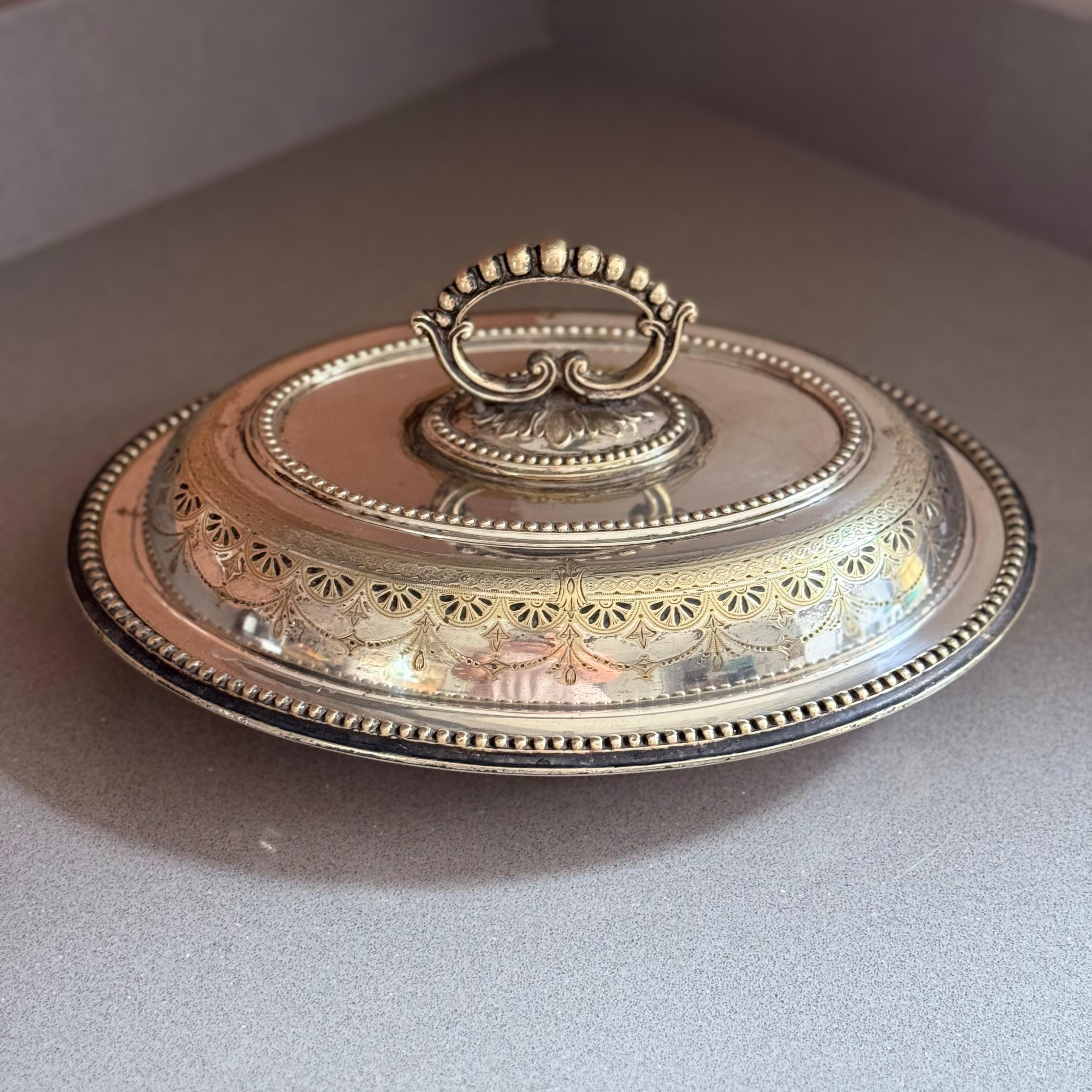 Cet élégant plat de service couvert ancien en métal argenté présente un décor gravé raffiné de style néoclassique, souligné par des motifs d'éventail et de guirlande festonnés autour du couvercle. Le bord perlé finement détaillé et la poignée