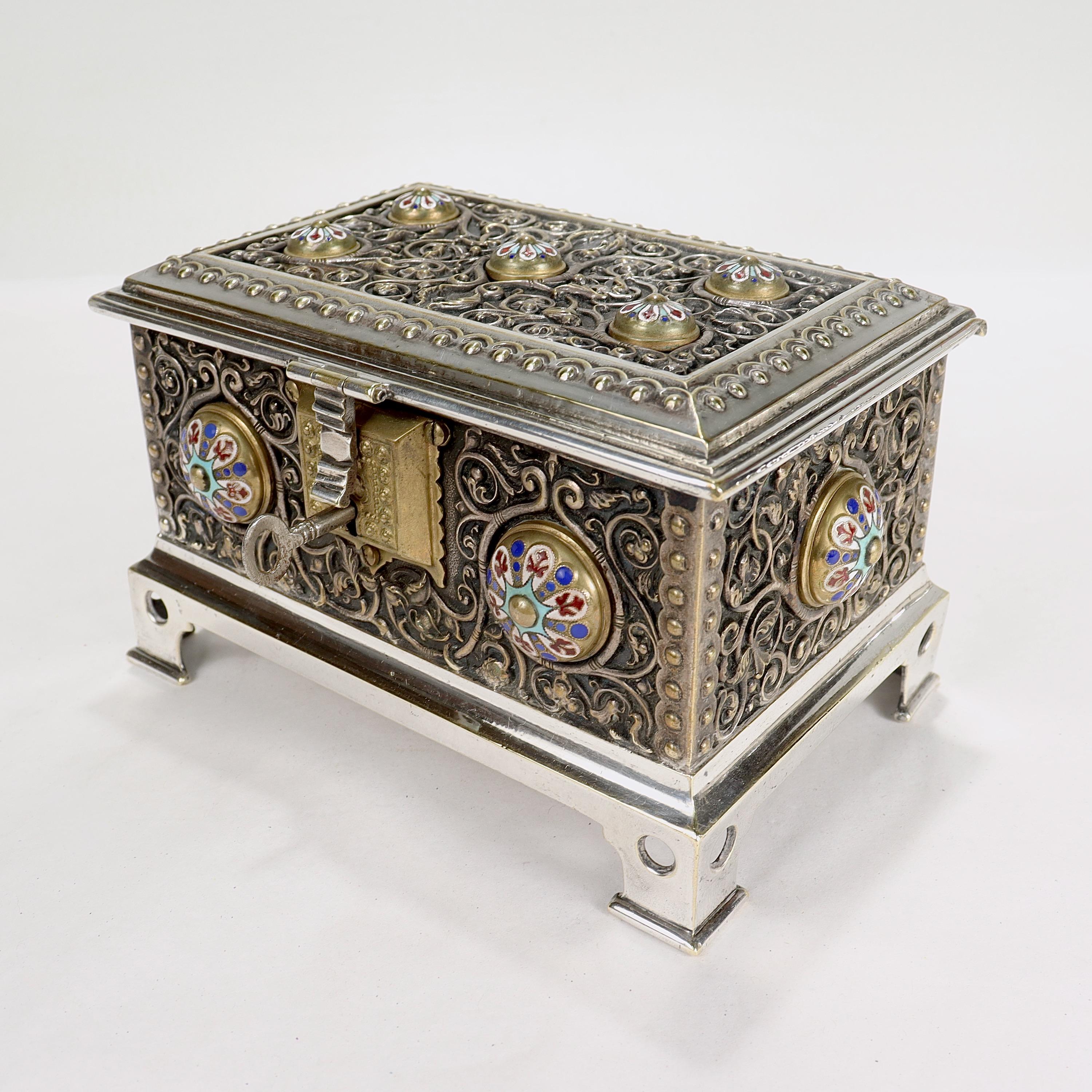 A fine silver & enamel table box or casket.

With stylized floral & vine work throughout, raised circular enameled cabochons on all sides, and raised bracket feet. 

The interior is lined with red velvet.

Marked with an M to the base and an 11 to