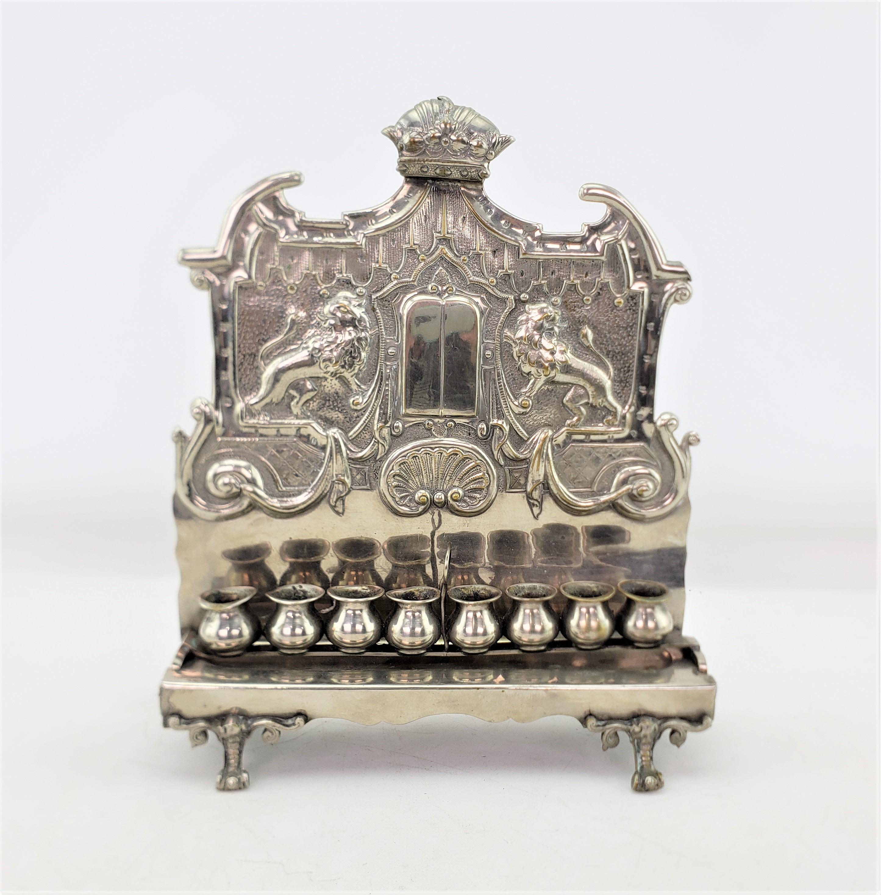 This antique menorah is unsigned, but presumed to have originated from Europe and date to approximately 1920 and done in a Victorian style. This menorah is composed of stamped metal with a silver platef finish with two rearing lions on the back