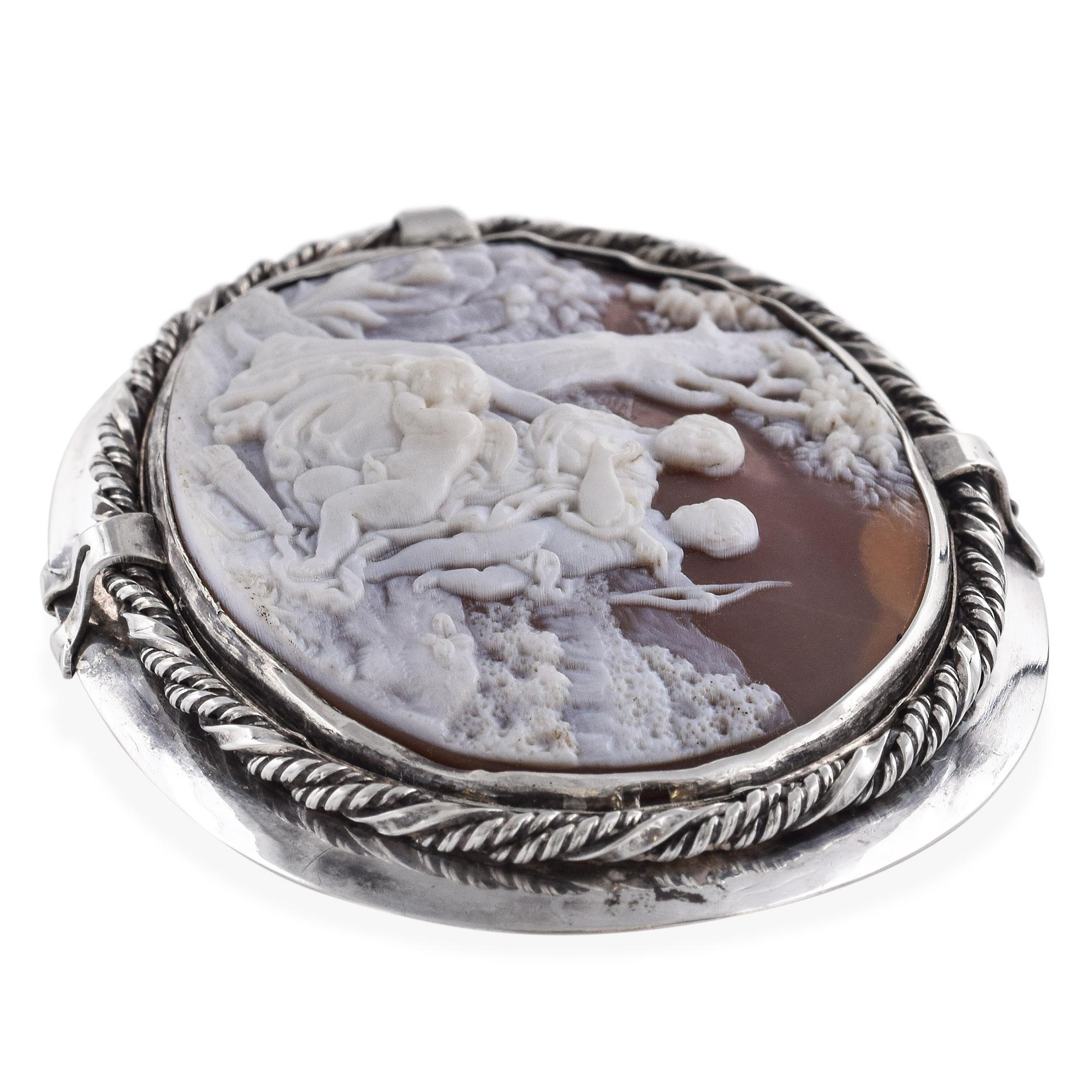 Este broche de plata antigua, bellamente detallado, presenta un camafeo de concha finamente tallado que representa a Venus, la diosa romana del amor, acompañada por Cupido y sus otros hijos querubines. La escena capta la gracia maternal y el afecto