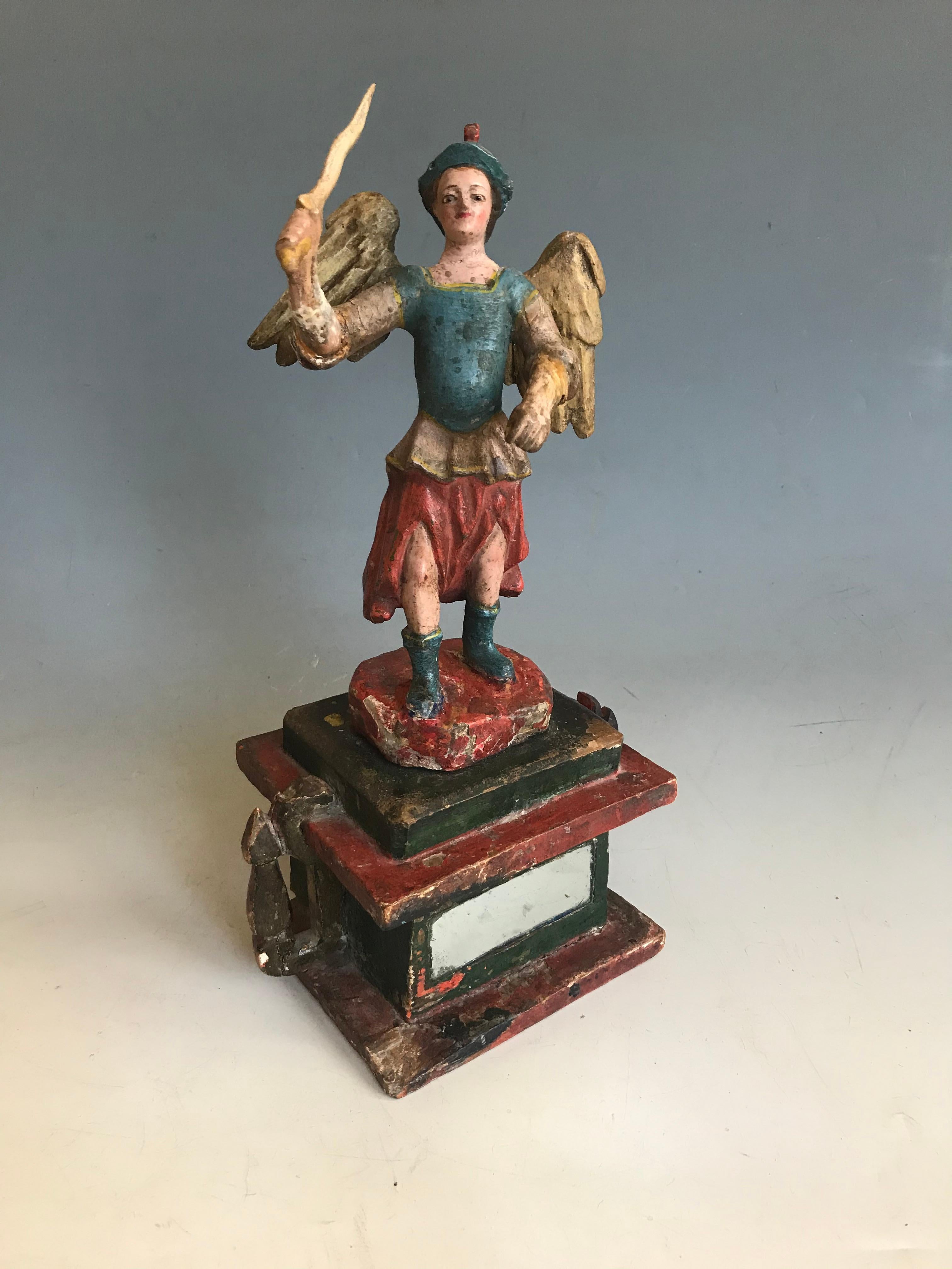 Antique South American Spanish Colonial Poly chrome Folk Art Santos figure of St Michael with sword. The finely modeled winged figure standing with a sword on a box type pedestal base with mirrored front, painted in red blue and cream, the face