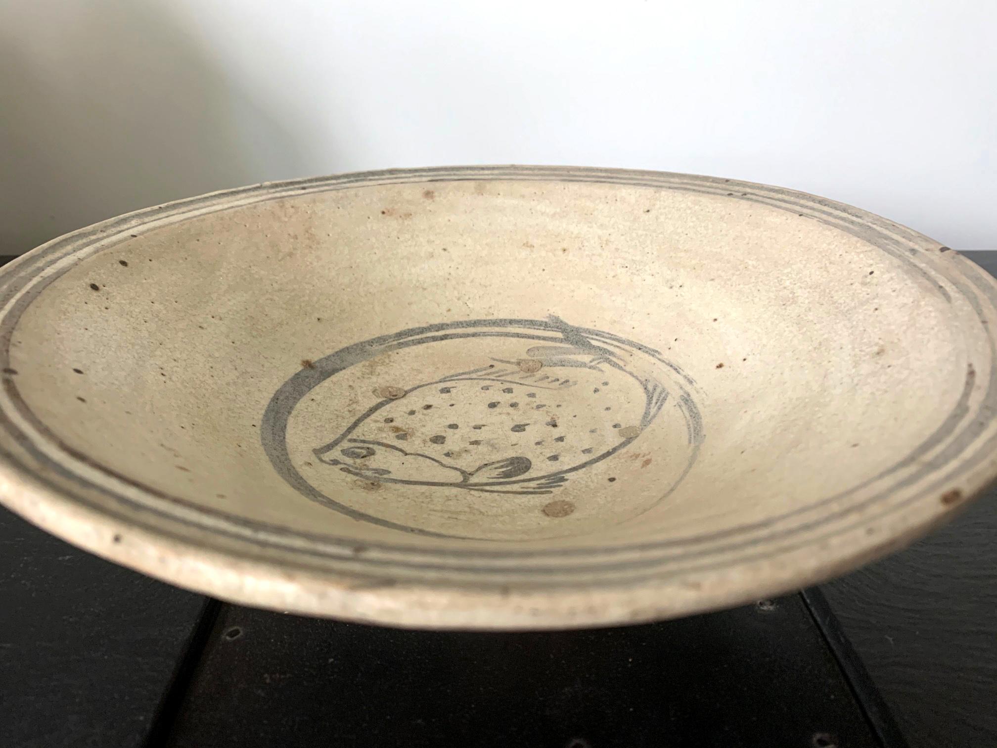 A shallow ceramic bowl from Sukhothai period of Thailand circa 14th-15th century. The dish was hand molded with a coarse pinkish clay and it has a white slip surface and a blue underglaze decoration. Three circular rings circle the rim and a double
