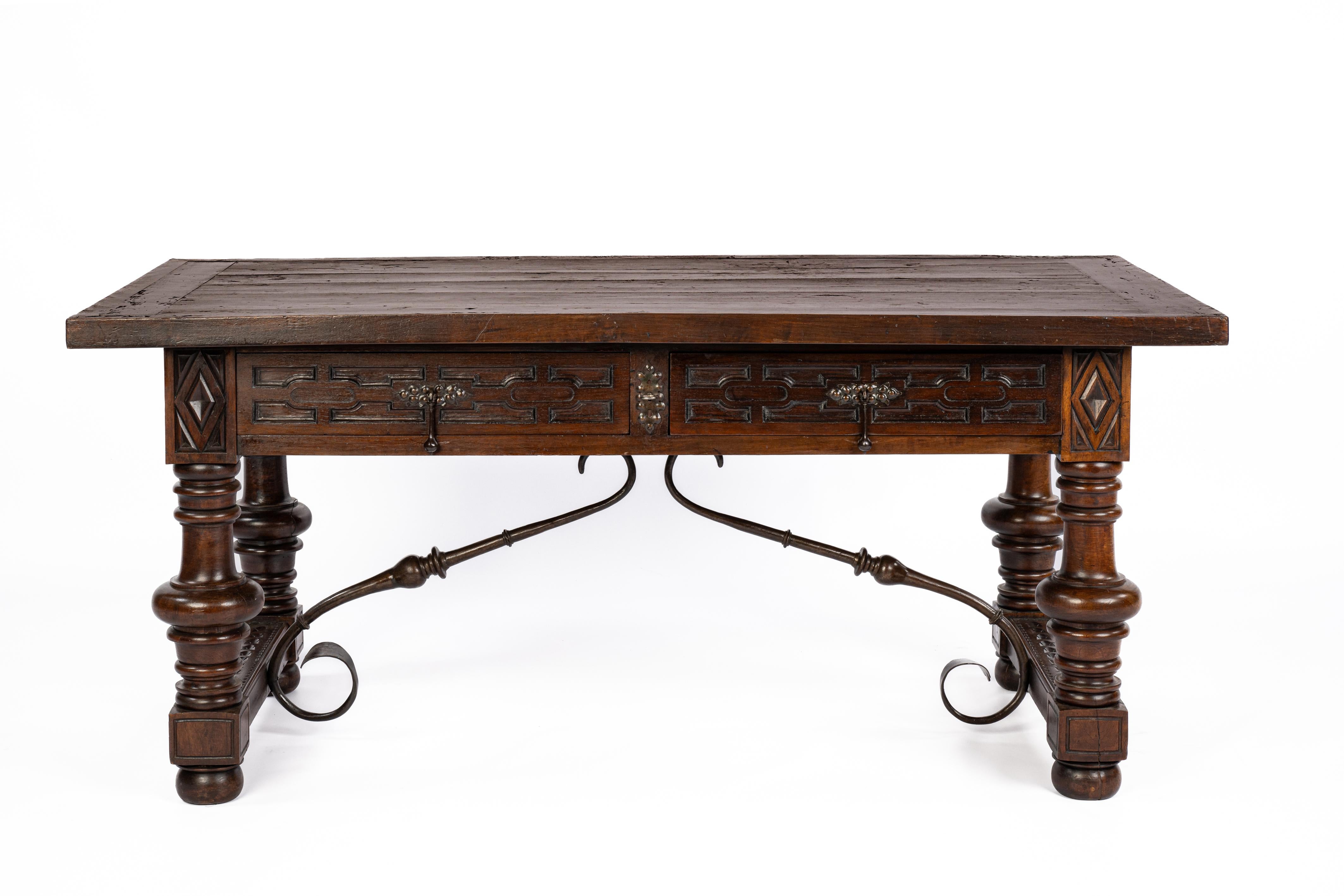 This beautiful antique late 19th-century Spanish Baroque desk or writing table is made of solid chestnut wood with bold-turned legs joined by wrought iron stretchers. The table features two drawers with a geometric carved front and forged steel drop