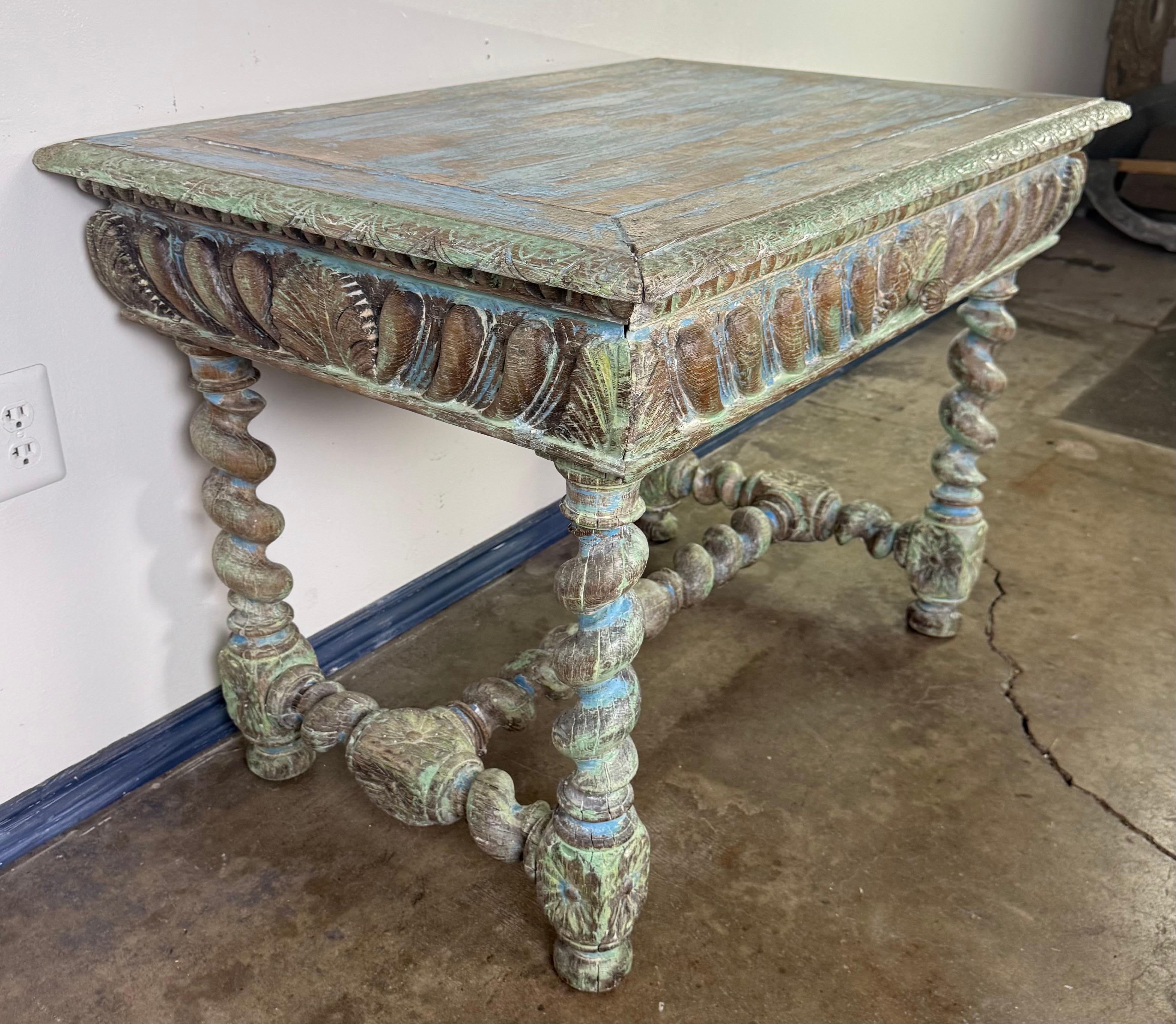 Una mesa de escritorio de estilo colonial español ricamente detallada, que muestra una magistral artesanía tallada a mano y una pátina pintada desgastada por el tiempo. La encimera rectangular presenta un borde biselado y descansa sobre un delantal