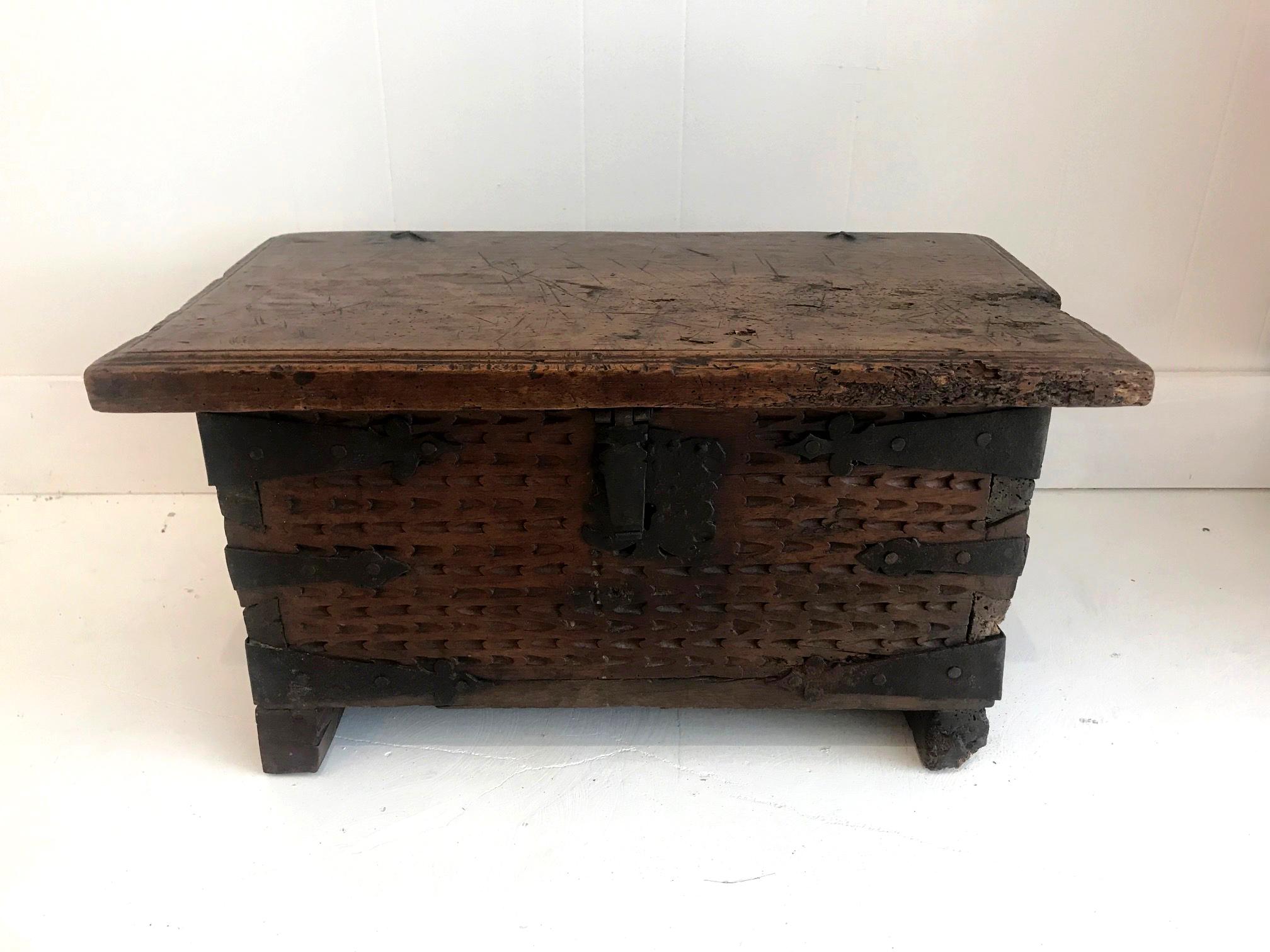 A small Spanish or Spanish Colonial trunk/coffer bench in carved walnut with large joinery, iron corner brackets and an iron strap lock, supported on a plinth base. The bench is likely dated to early 19th century. The carving was hand-chiselled in a