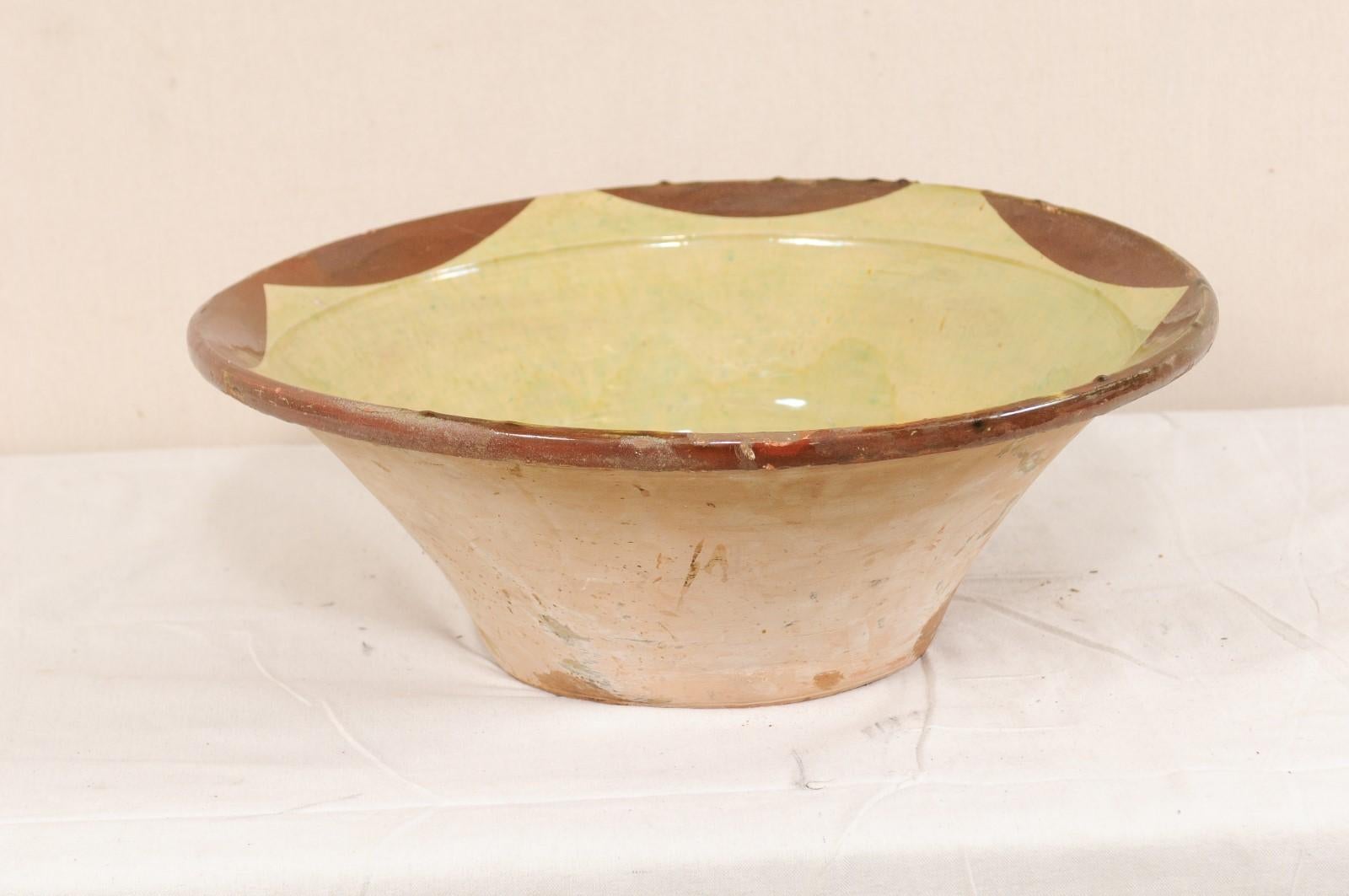 A Spanish terracotta bowl with pale green and brown accents from the early 20th century. This antique bowl from Spain is round in shape, with widest part being at the top lip, it gradually slopes inward towards the center, and rests upon a flat