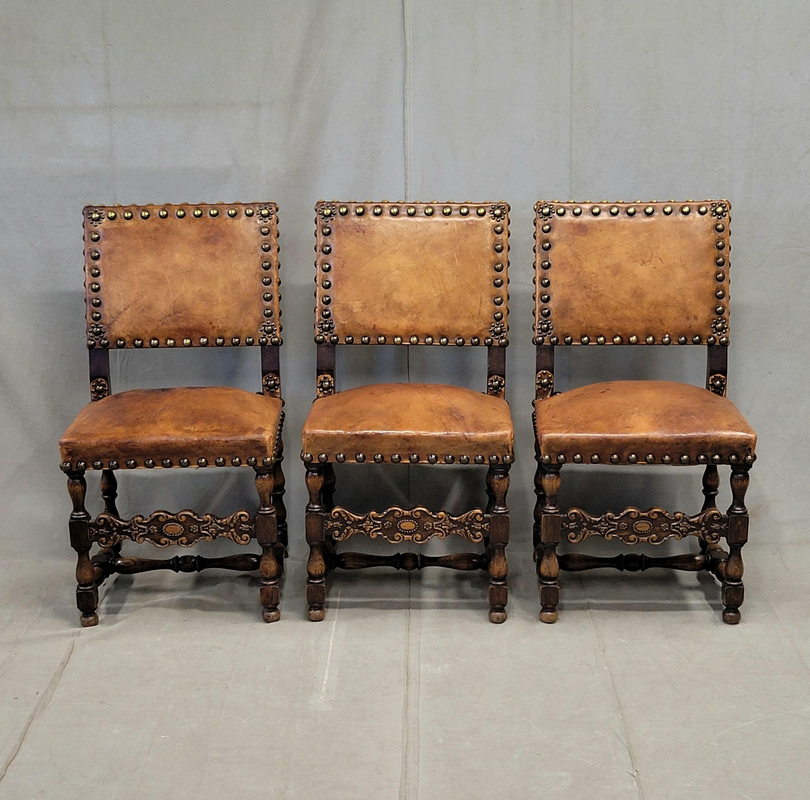 A classic set of three rustic European revival chairs, likely from Spain or Italy. The oak frames are strong and stable. Note the intricately carved oak stretchers across the front. The original oversized brass tacks are all intact and add such