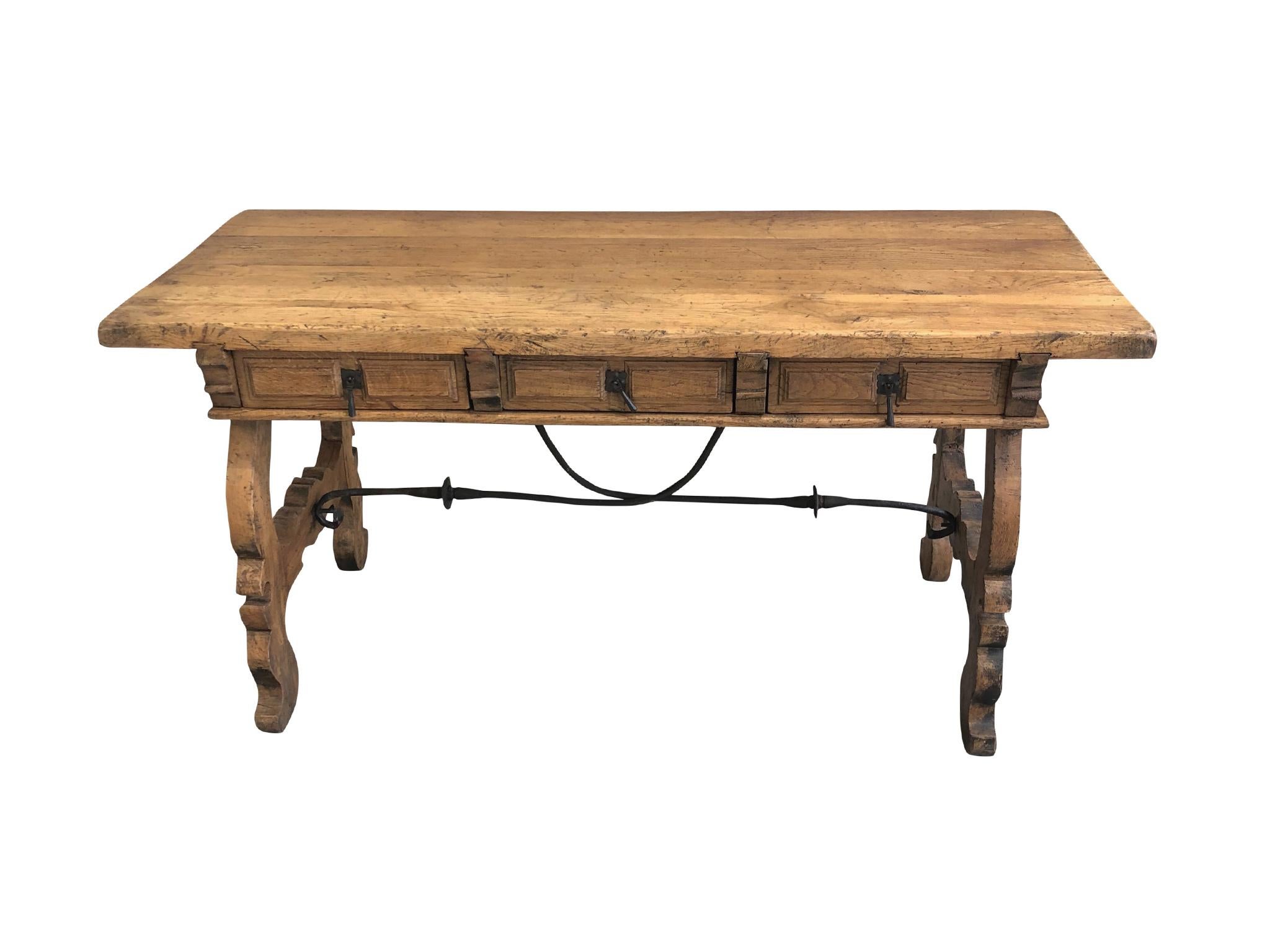 An antique desk handcrafted in a Spanish Colonial style. Made in the 19th century. The desk consists of carved and hand-joined oak wood. There are three drawers with iron pulls. A wrought iron bar supports the trestle legs. The beautifully aged and