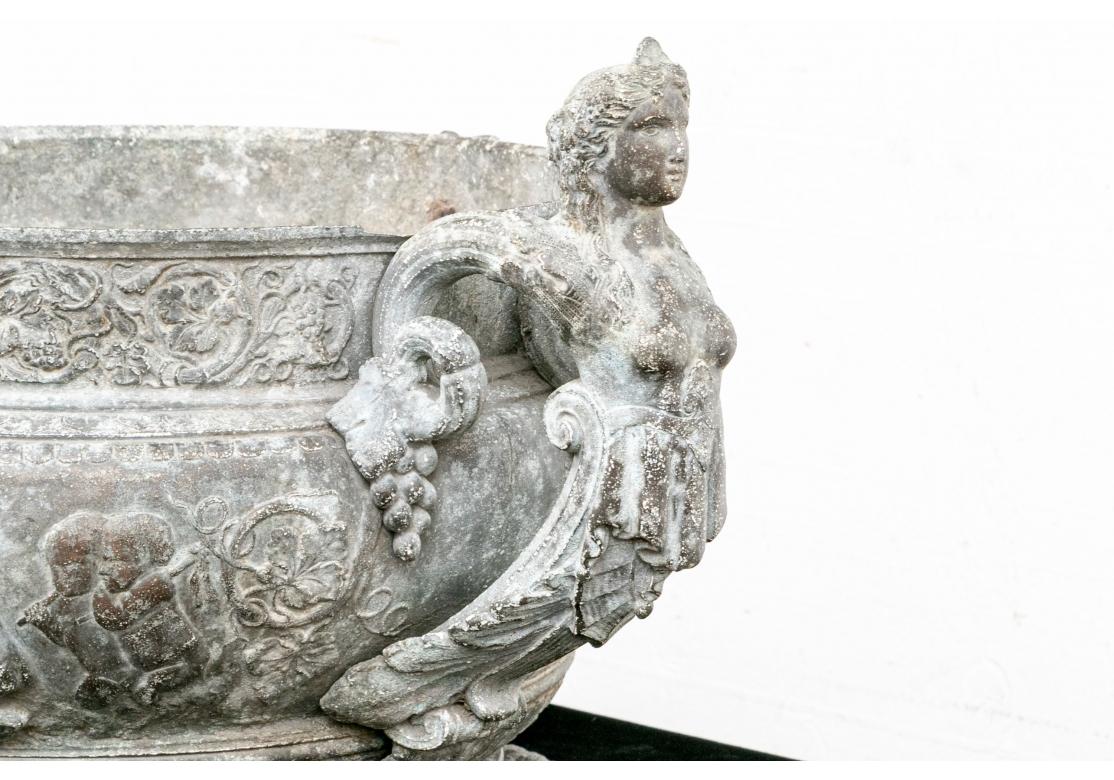 The basin with decorative bands in relief with grape vines and a wide band of puttii at their tasks in Bacchic revelry. Masks of Pan in relief on the top band. With semi nude female figural handles ending in leaves and scrolled grape vines. A