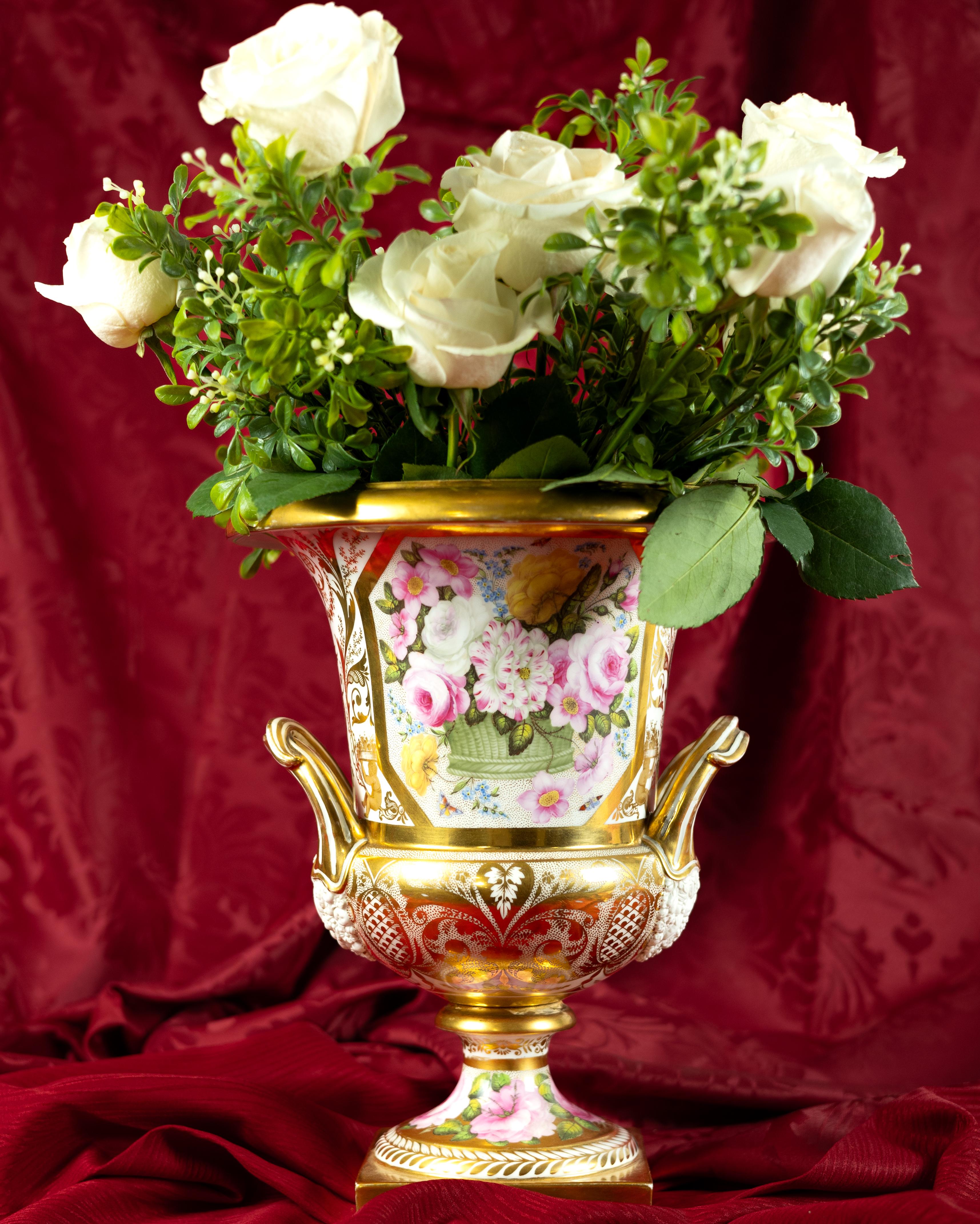  This significant Regency period campana-shaped urn is finely painted with fabulous pink and yellow roses and small blue forget-me-nots overflowing from a green basket. 
The reverse shows beautiful pink roses (see image #2).  
The elaborate and