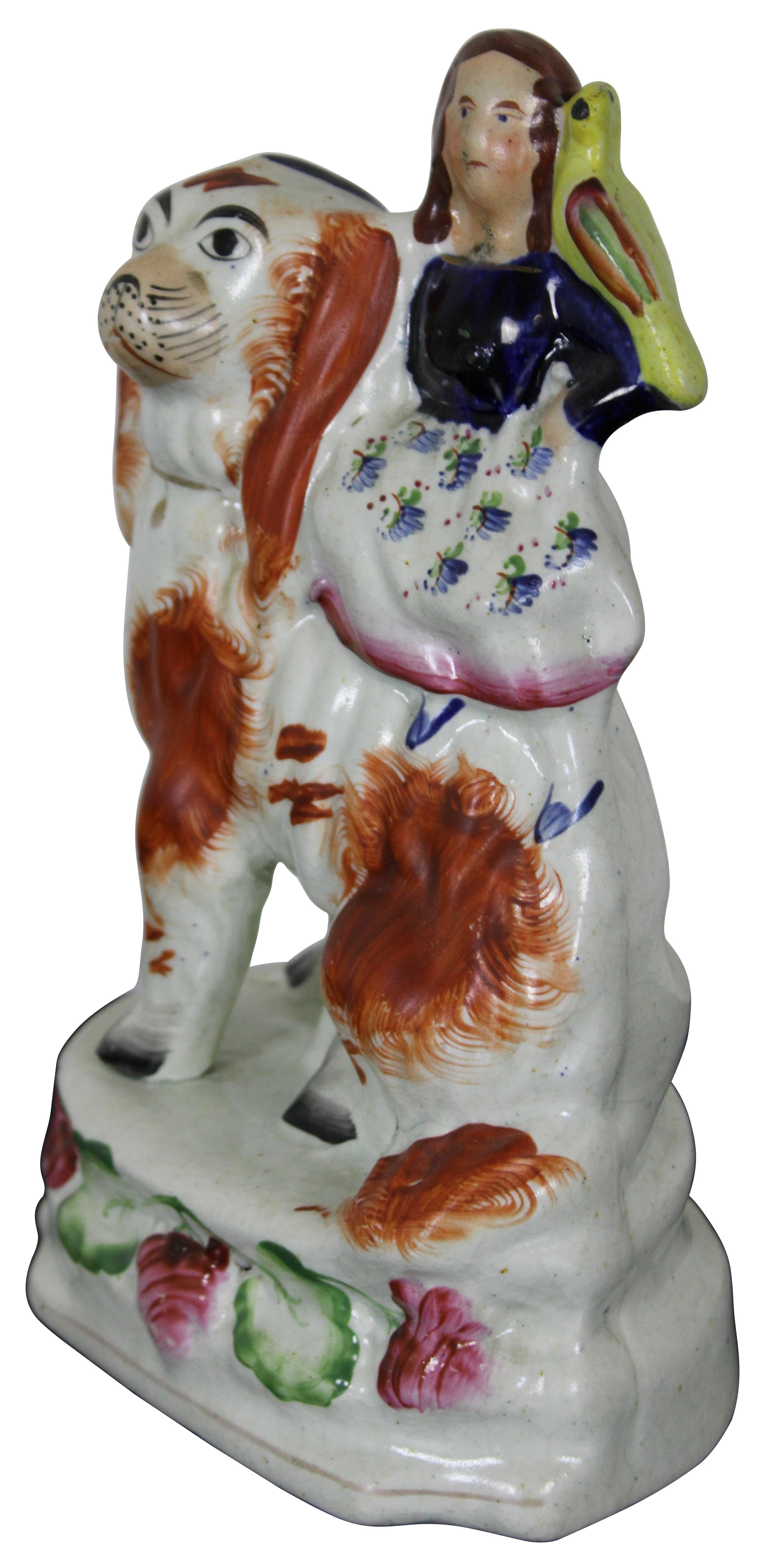 Antique 19th century Staffordshire porcelain figurine featuring a small girl with a yellow bird on her shoulder, seated on an oversized red and white King Charles Spaniel, mounted on a floral base. Measure: 8