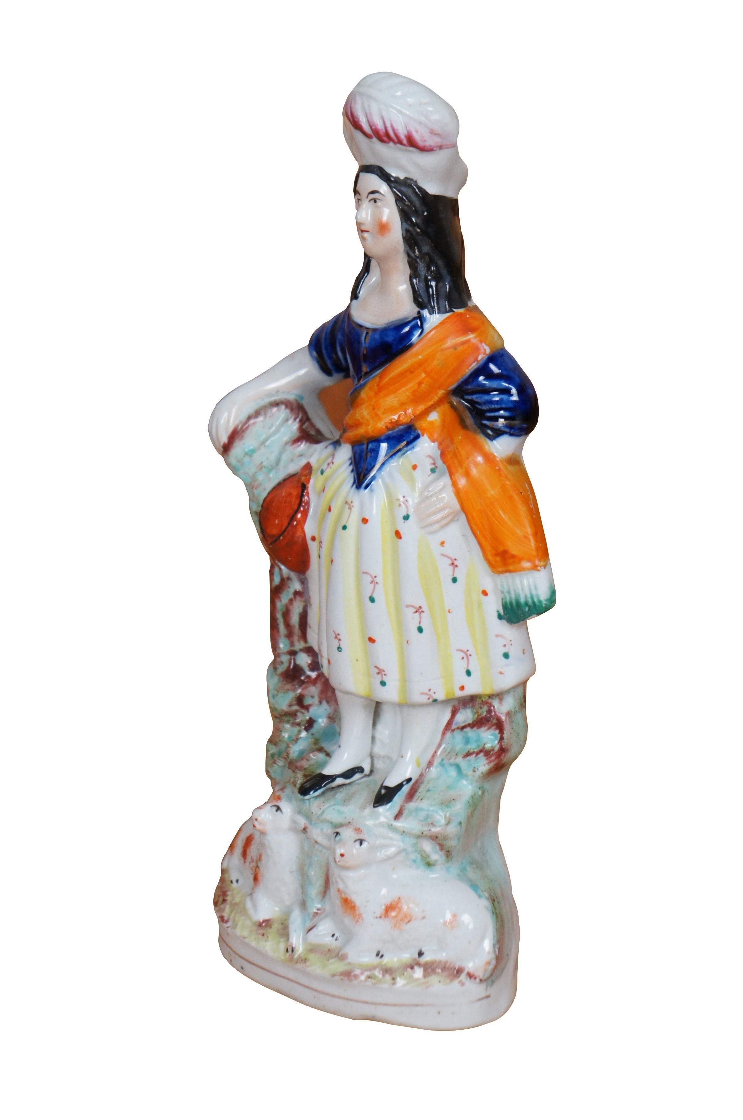 Antique 19th century Staffordshire porcelain flatback figurine depicting a s Scottish shepherdess in feathered hat, cobalt blue jacket, orange erisade / scarf, and patterned yellow skirt, resting one hand on a mossy tree stump with two lambs / sheep