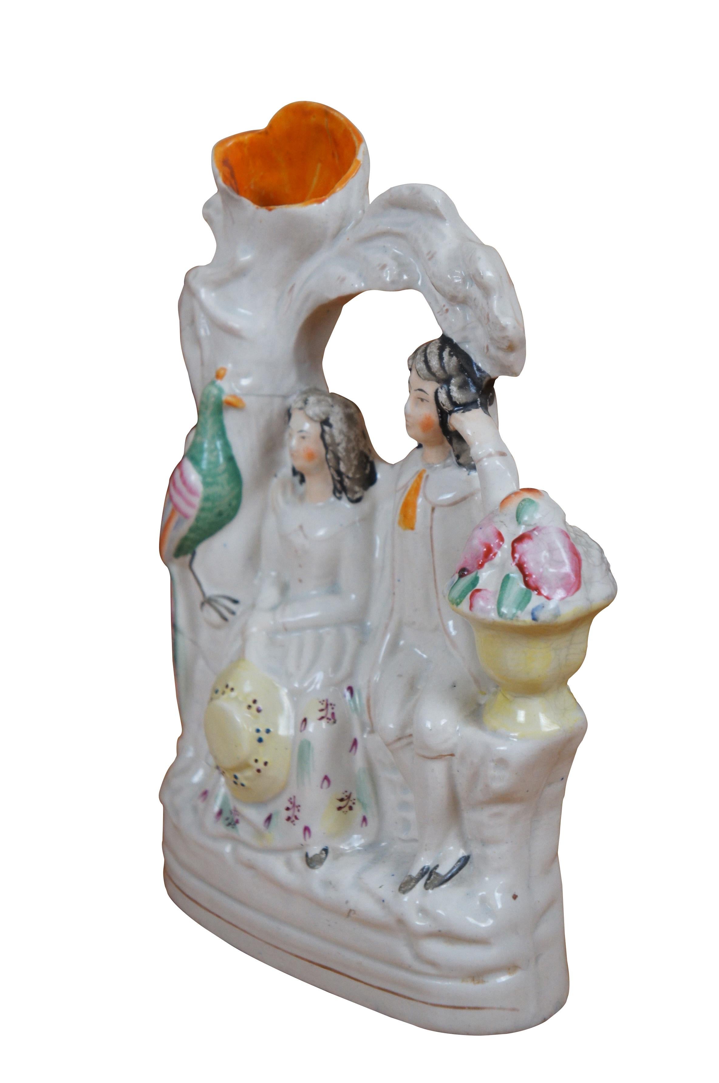 Antico vaso da versare in porcellana inglese dello Staffordshire del XIX secolo, a forma di coppia innamorata e corteggiata seduta su un muro del giardino sotto un albero, affiancata da un'urna di fiori e da un fagiano/pappagallo/uccello dai colori