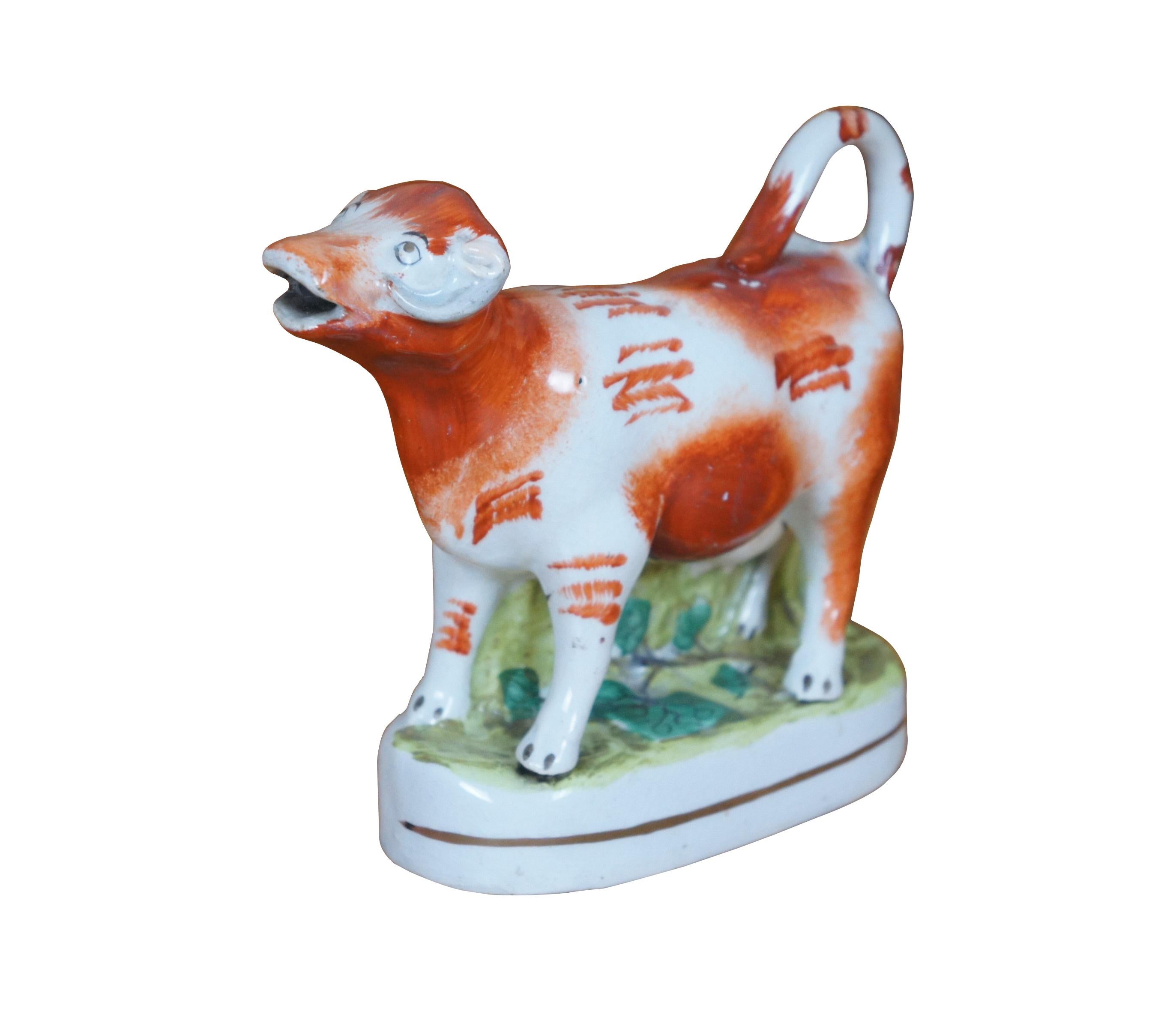 Antique 19th century Staffordshire porcelain flatback figural cream pitcher / bud vase in the shape of a red and white cow next to a tree stump, on a grassy oval base. Base painted with grass and English ivy. Tail curls to form