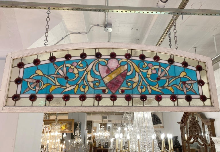 Early 1900s arched stained glass transom window. The wide width and highly ornate design shows this was part of an over the top entranceway. The large center shield is flanked by smaller jewels in vivid red, yellow and blue colors. This can be seen