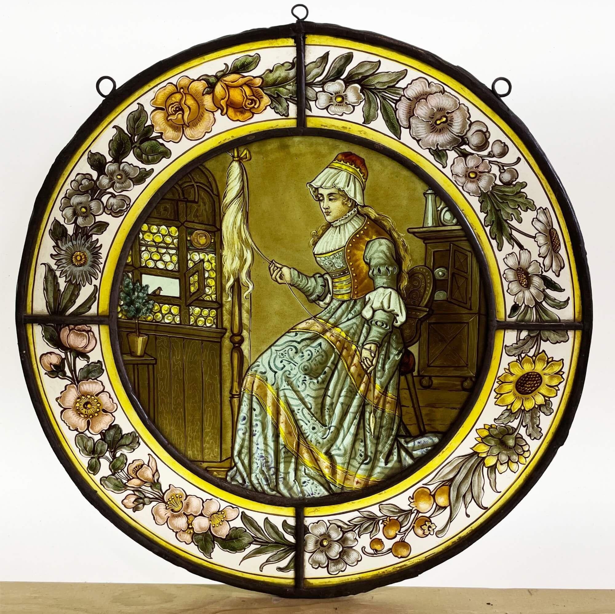An antique stained glass window depicting a Victorian woman spinning yarn. Hand-painted stylised flowers and a vibrant yellow border surround the woman while she effortlessly spins the yarn. Her dress is a pale blue and gold displaying wealth and